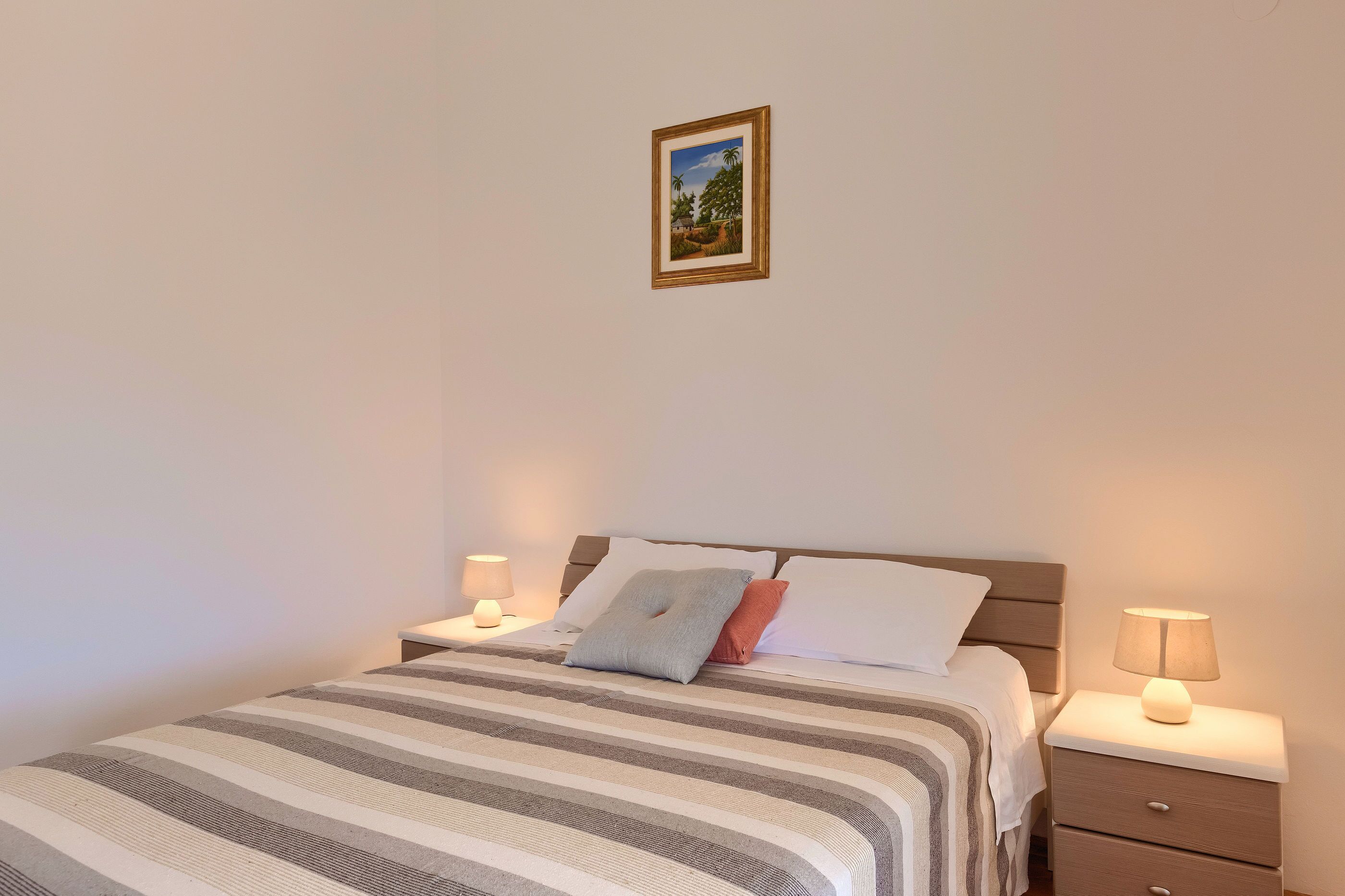 Una cama bien hecha con ropa de cama a rayas, dos mesitas de noche con lámparas y un cuadro en la pared.