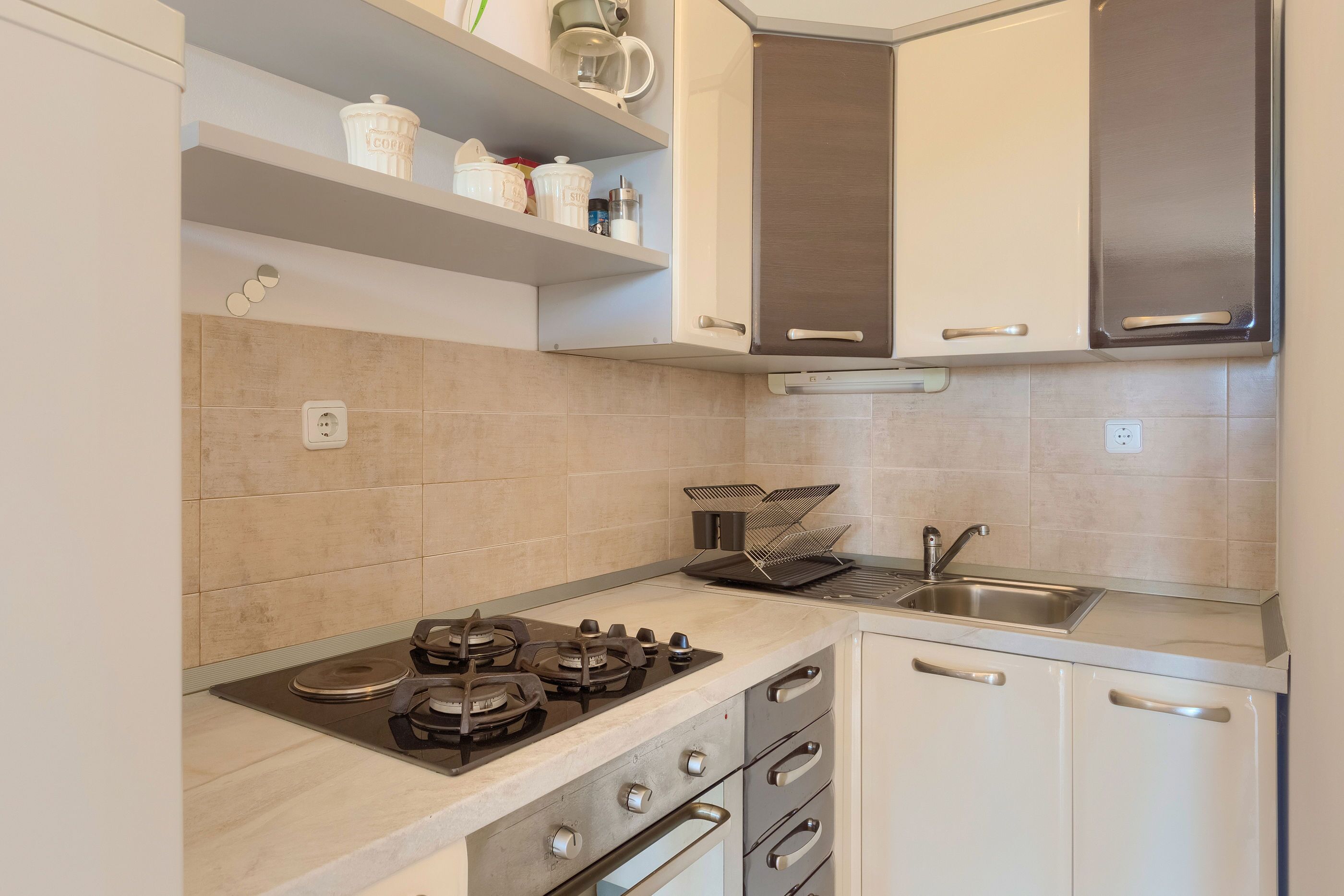 Una cocina moderna con estufa a gas, horno y pileta de acero inoxidable. Hay armarios blancos y una encimera con una cafetera y otros utensilios de cocina.