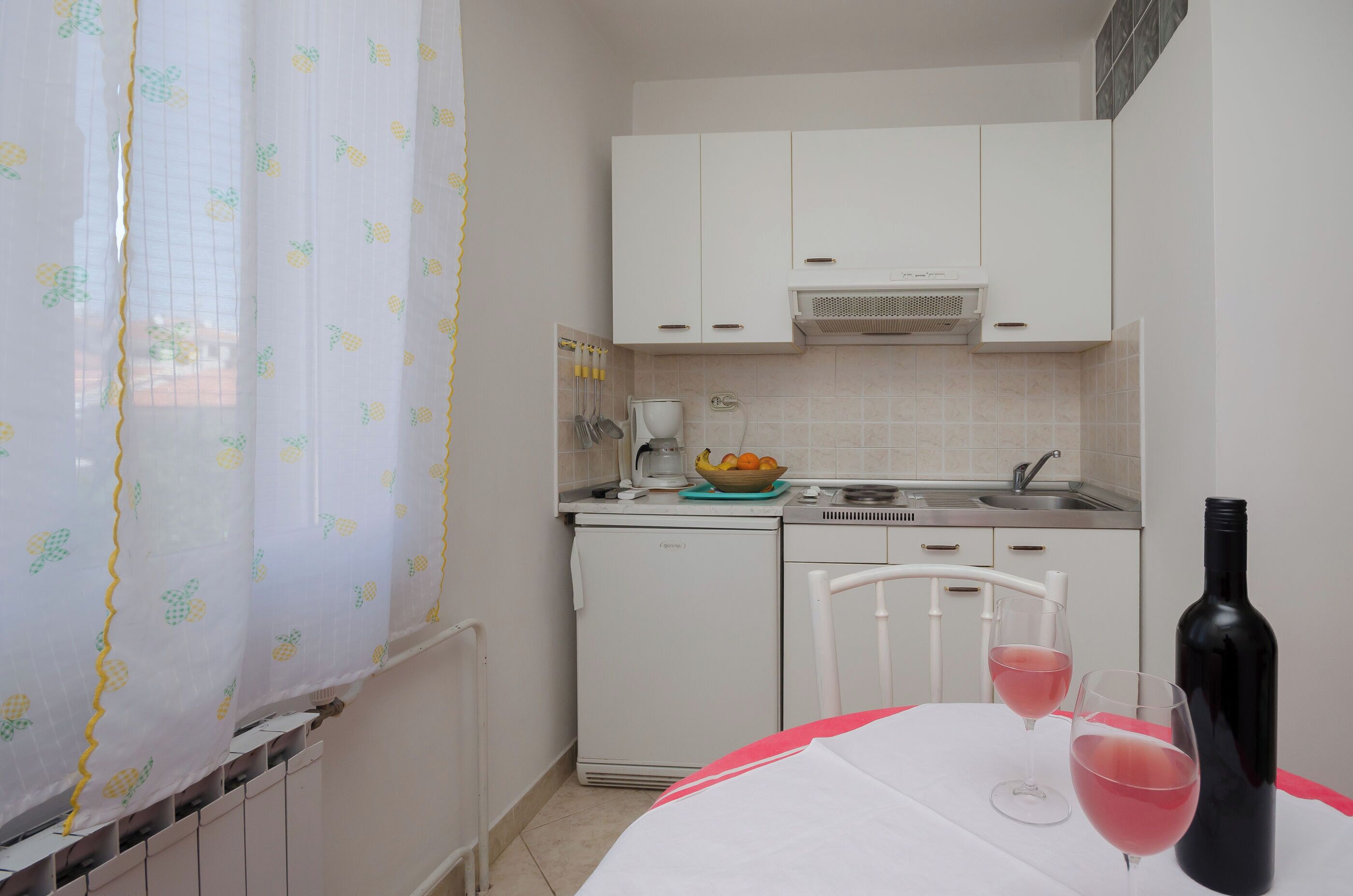 A compact kitchen with white cabinets, a small refrigerator, and a table with two glasses of red wine and a bottle.