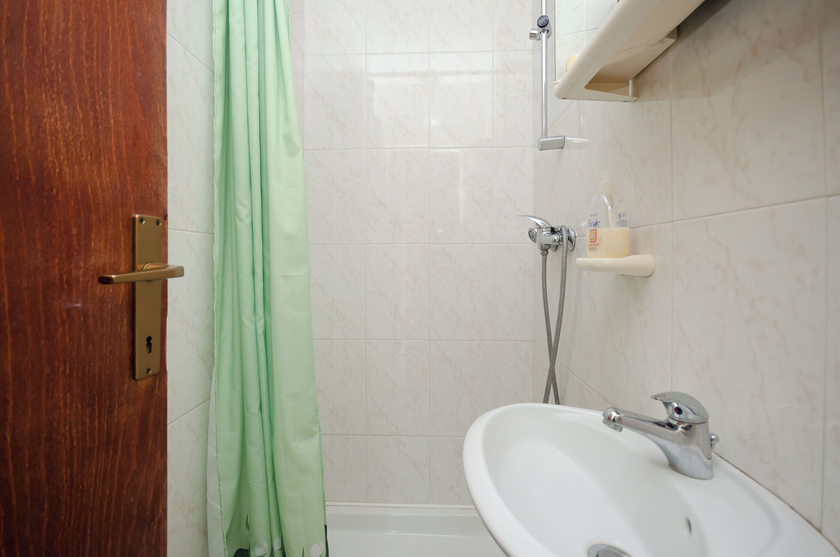 Une salle de bain avec une porte en bois, un rideau de douche vert, un lavabo blanc et une douche équipée d’un pommeau de douche à main.