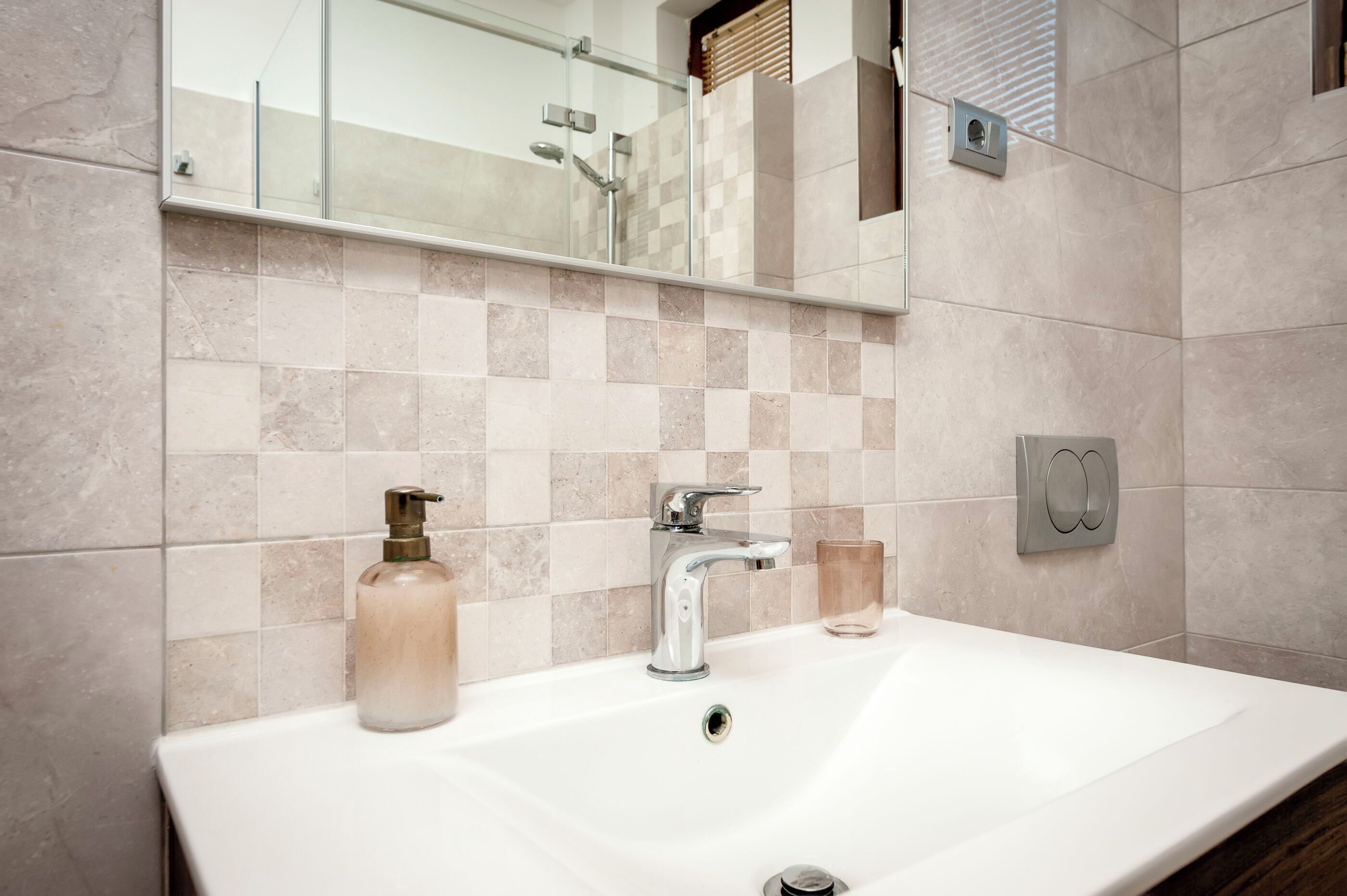 A bathroom with a white sink, a mirror, a glass shower enclosure, and beige tiled walls.