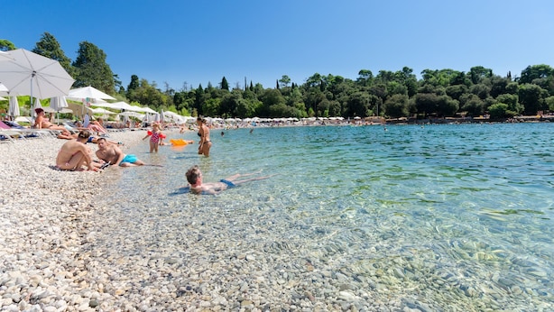 En strand med småsten, klart vatten, parasoller och folk som solar.