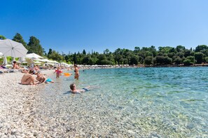 Ein Kiesstrand mit klarem Wasser, Sonnenschirmen und badenden Menschen.