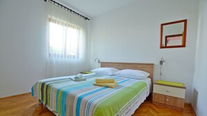 Ein ordentlich bezogenes Bett mit einer gestreiften Tagesdecke, ein Nachttisch mit einer Lampe und ein Fenster mit transparenten Vorhängen.