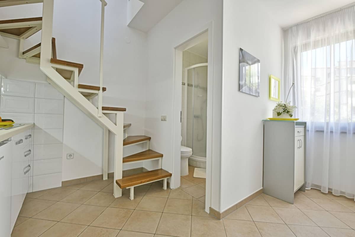 A modern kitchen with white cabinets, a wooden staircase, a toilet, and a small cabinet with a vase of flowers.