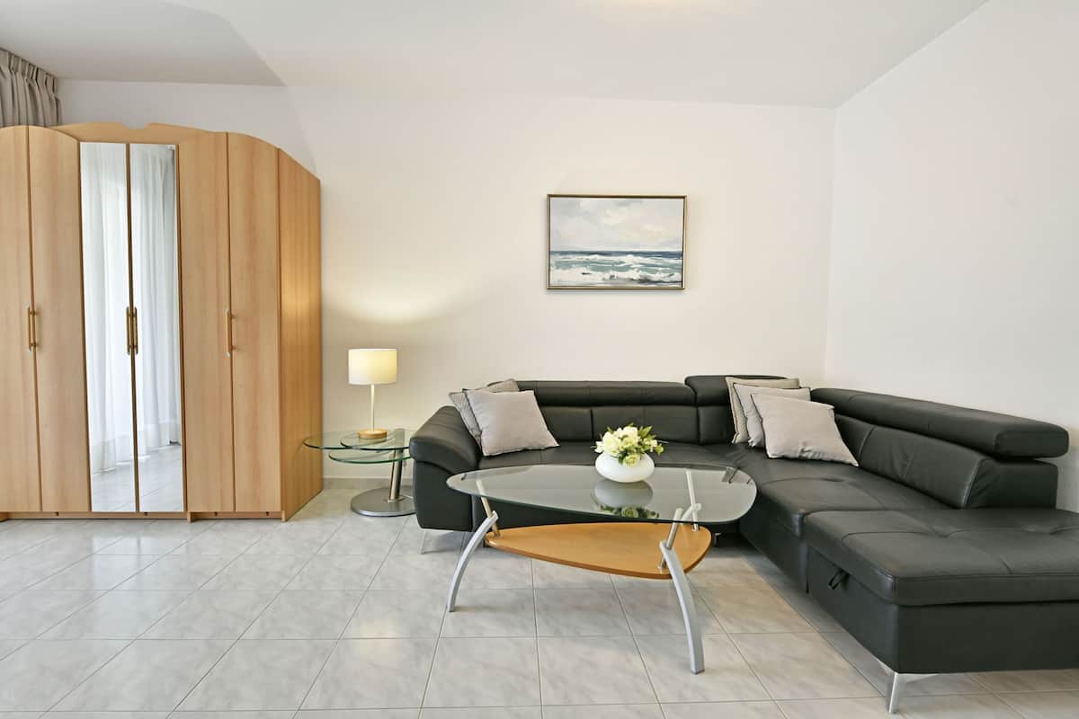A modern living room with a black leather sofa, a glass coffee table with a wooden base, a wooden wardrobe, a small side table with a lamp, and a framed picture on the wall.