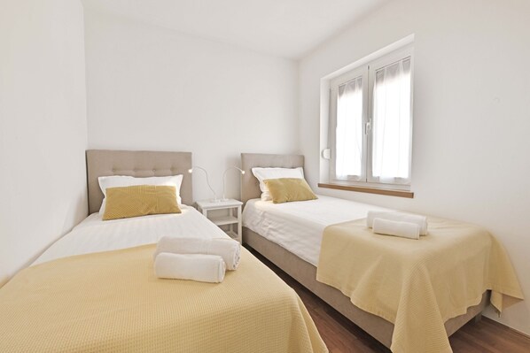 A hotel room with two single beds, each with a beige headboard, white bedding, and a yellow throw blanket. There is a nightstand with a lamp and a window with curtains.