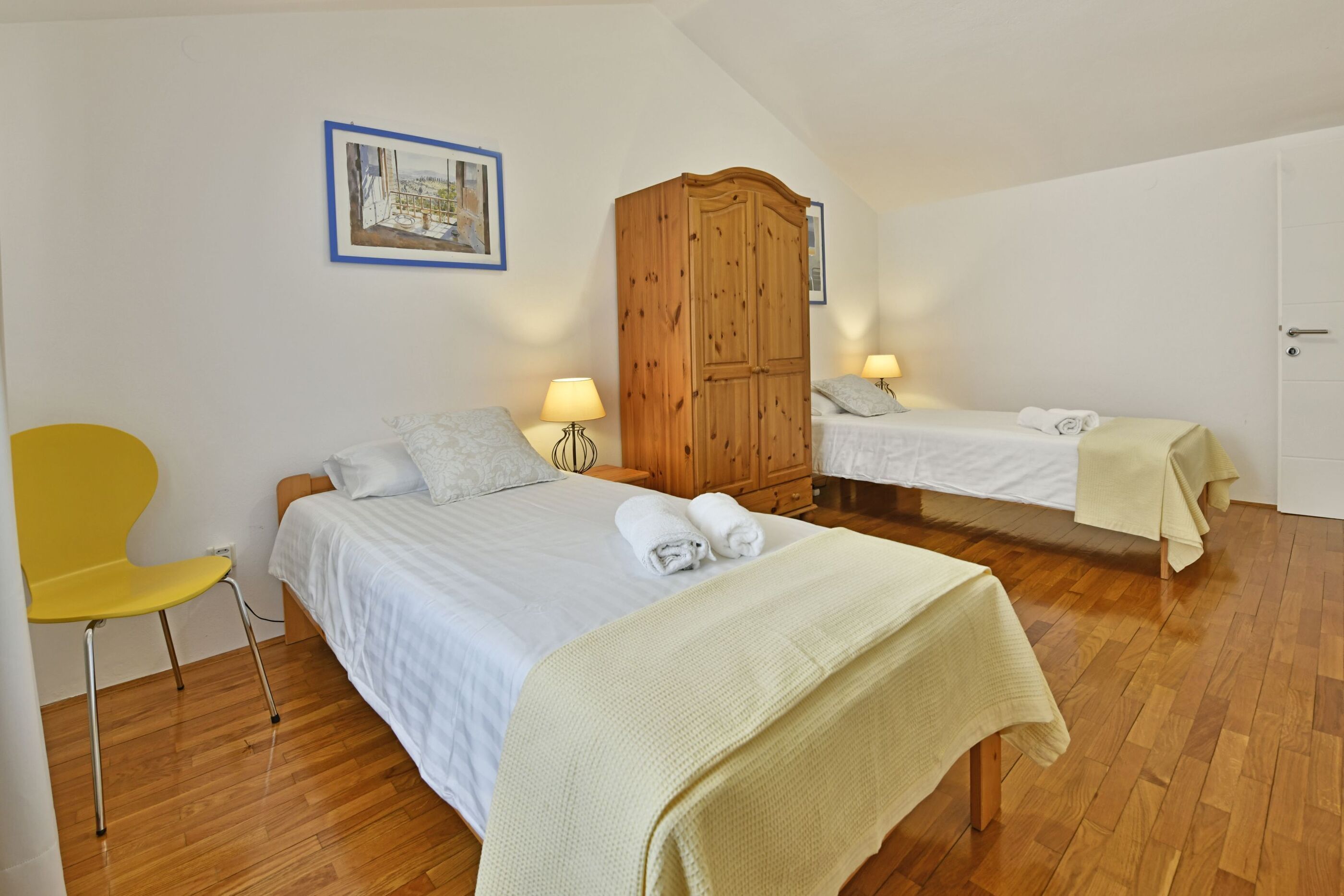 A bedroom with two beds, a wooden wardrobe, a yellow chair, and a framed picture on the wall.
