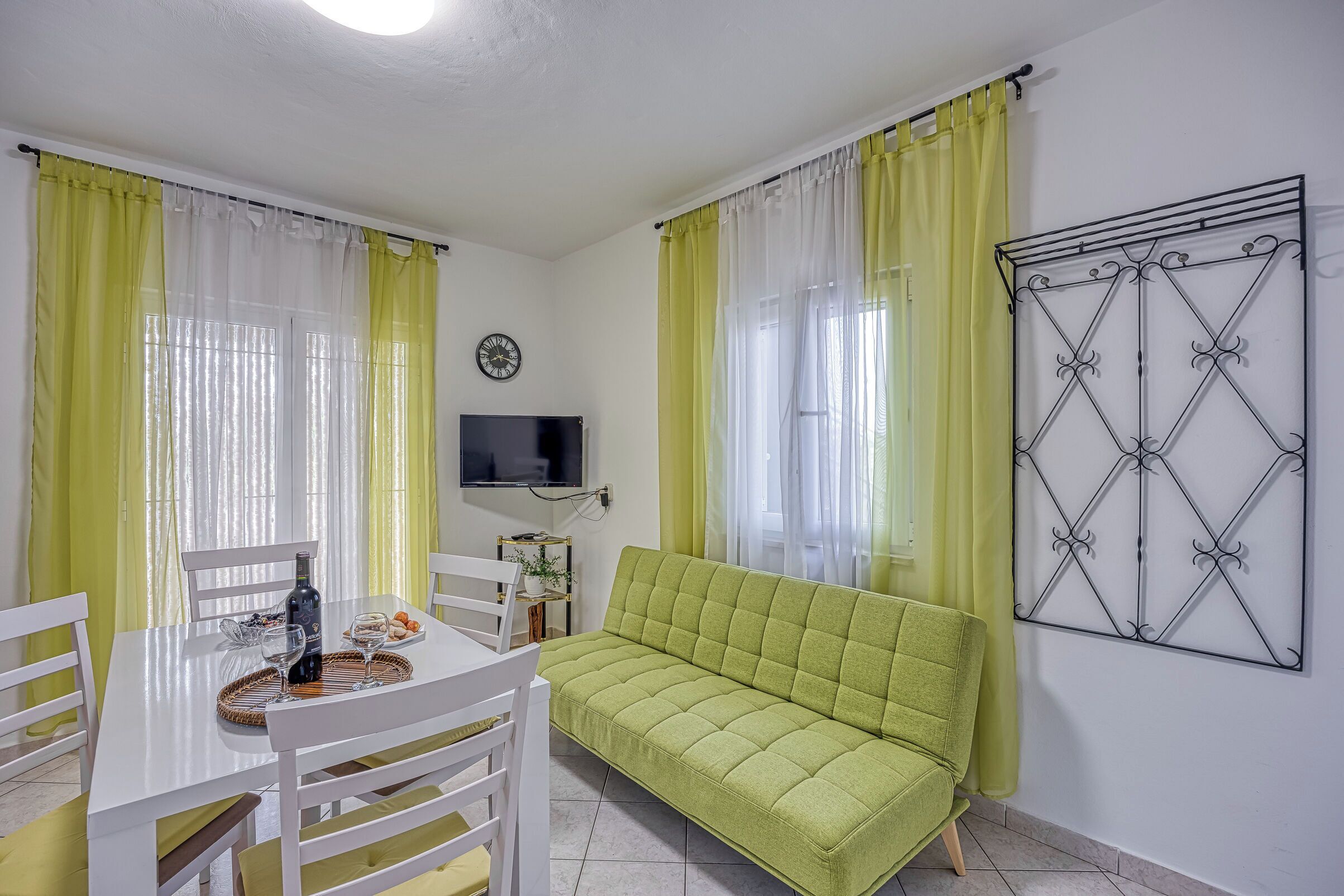 A room with a green sofa, white chairs, a dining table with a bottle and glasses, yellow curtains, and a TV on the wall.