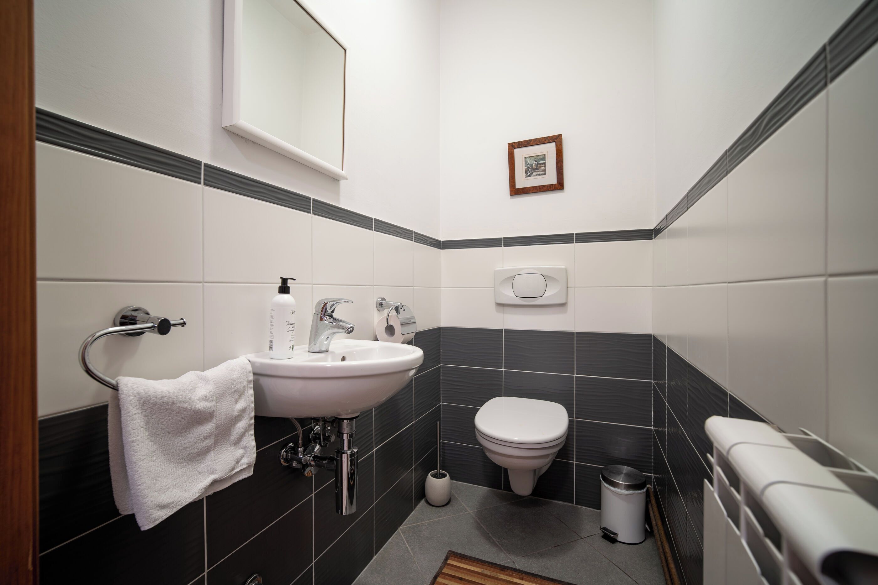 A modern bathroom with a white sink, black tiled walls, a toilet, and a trash bin.