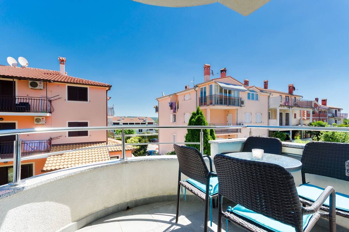 A balcony with wicker chairs and a table, overlooking a row of pastel-colored buildings.
