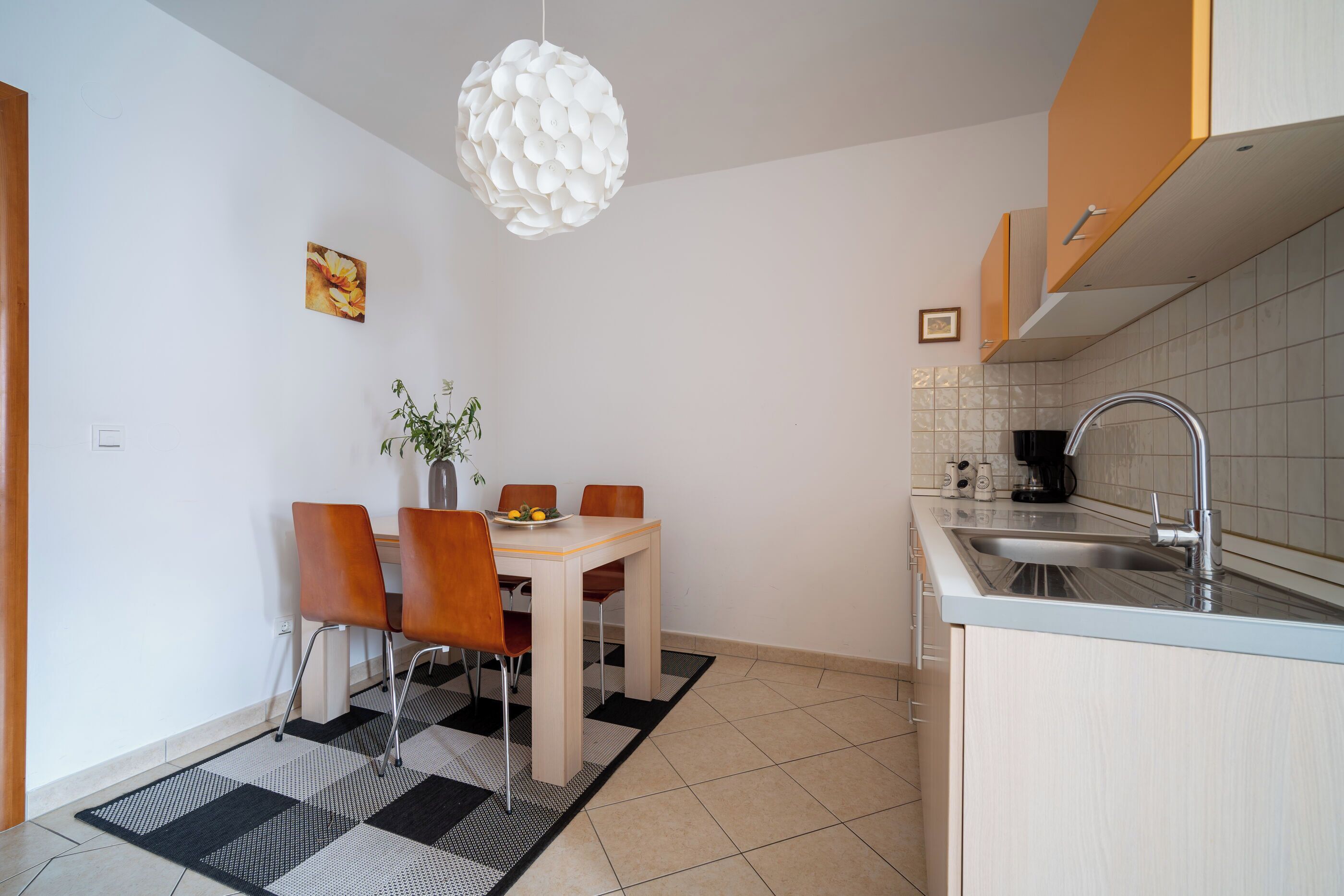 A compact kitchen with a dining area, featuring a modern pendant light, a small dining table with chairs, a sink, and a tiled backsplash.