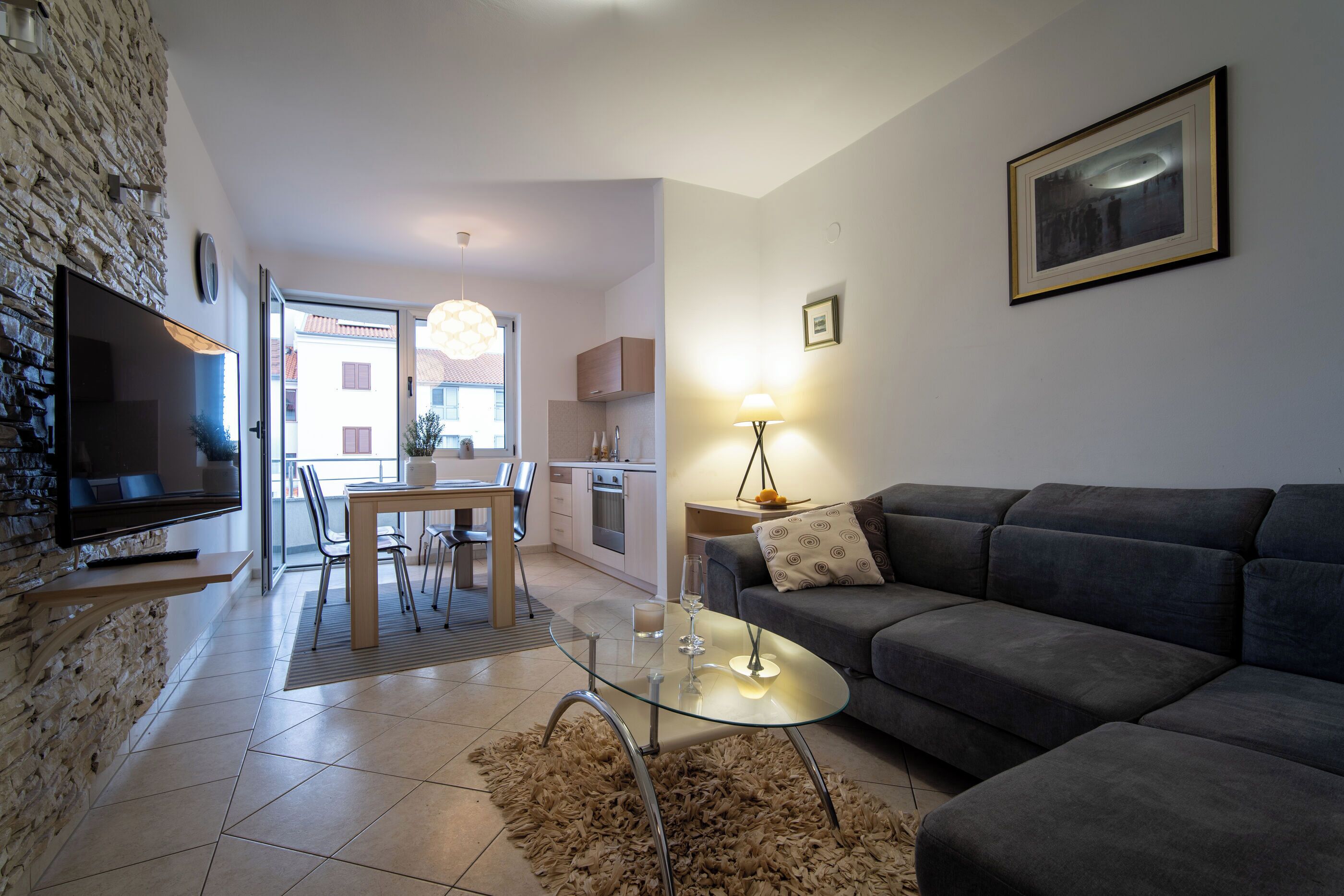Una sala moderna con un televisor de pantalla plana, un sofá gris, una mesa de centro de vidrio y una zona de comedor con una mesa y sillas de madera.