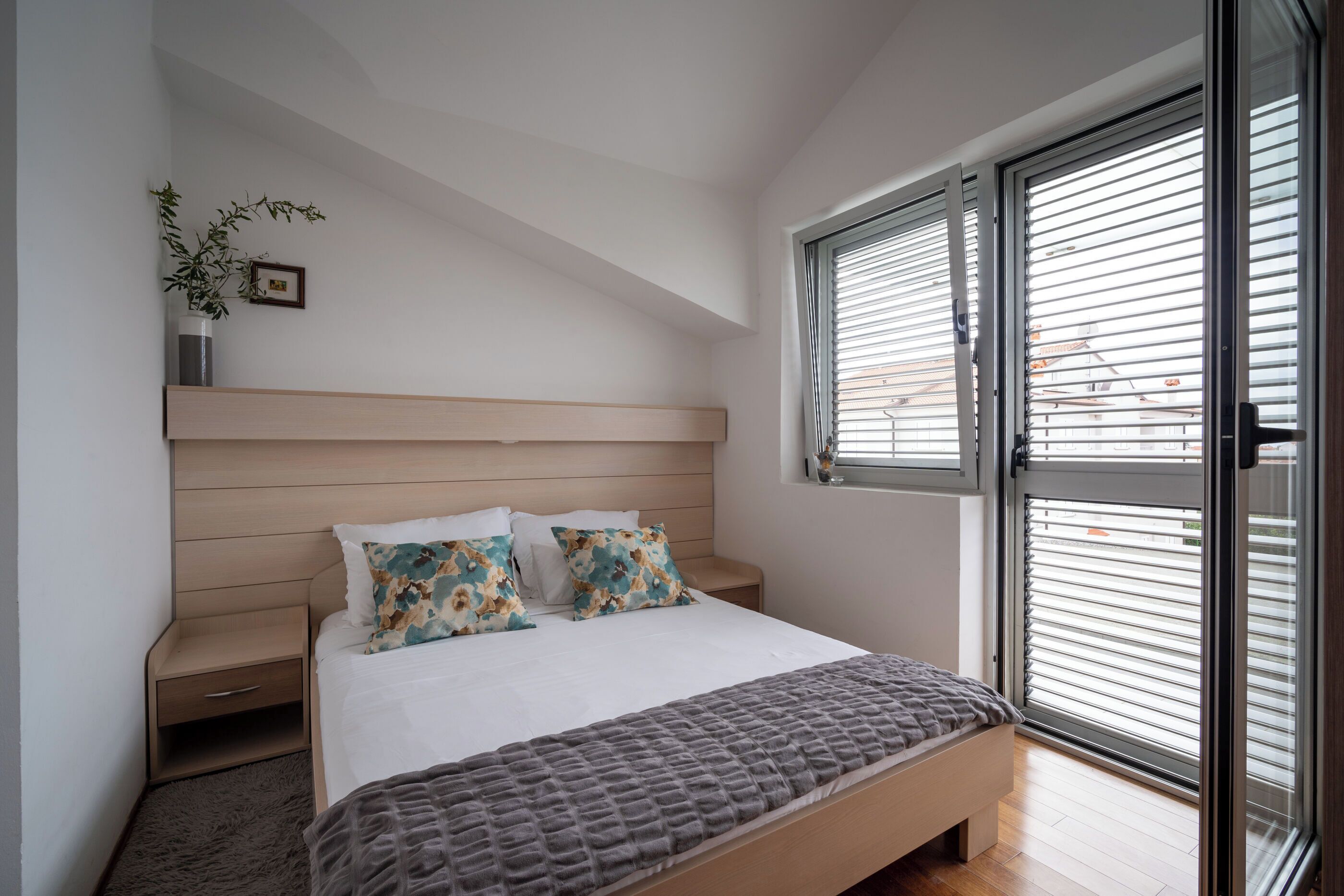 Un dormitorio con una cama, mesitas de noche, una planta en la pared y una ventana con persianas.