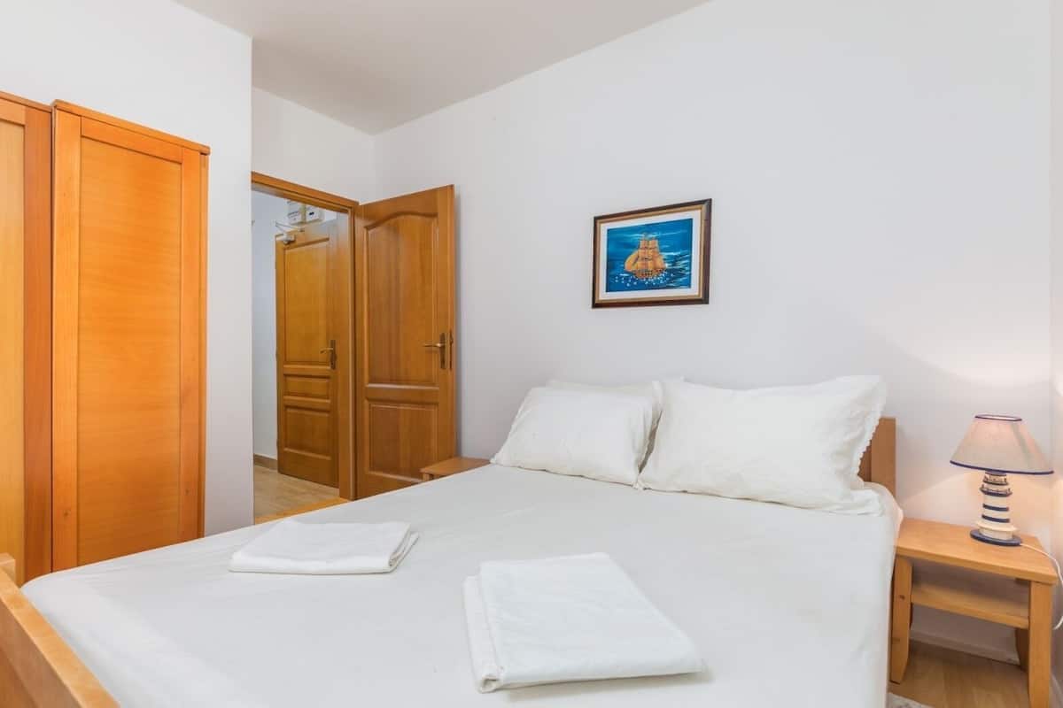 Une chambre à coucher avec un lit, du linge de lit blanc, une armoire en bois, une table de chevet avec une lampe et un tableau encadré au mur.