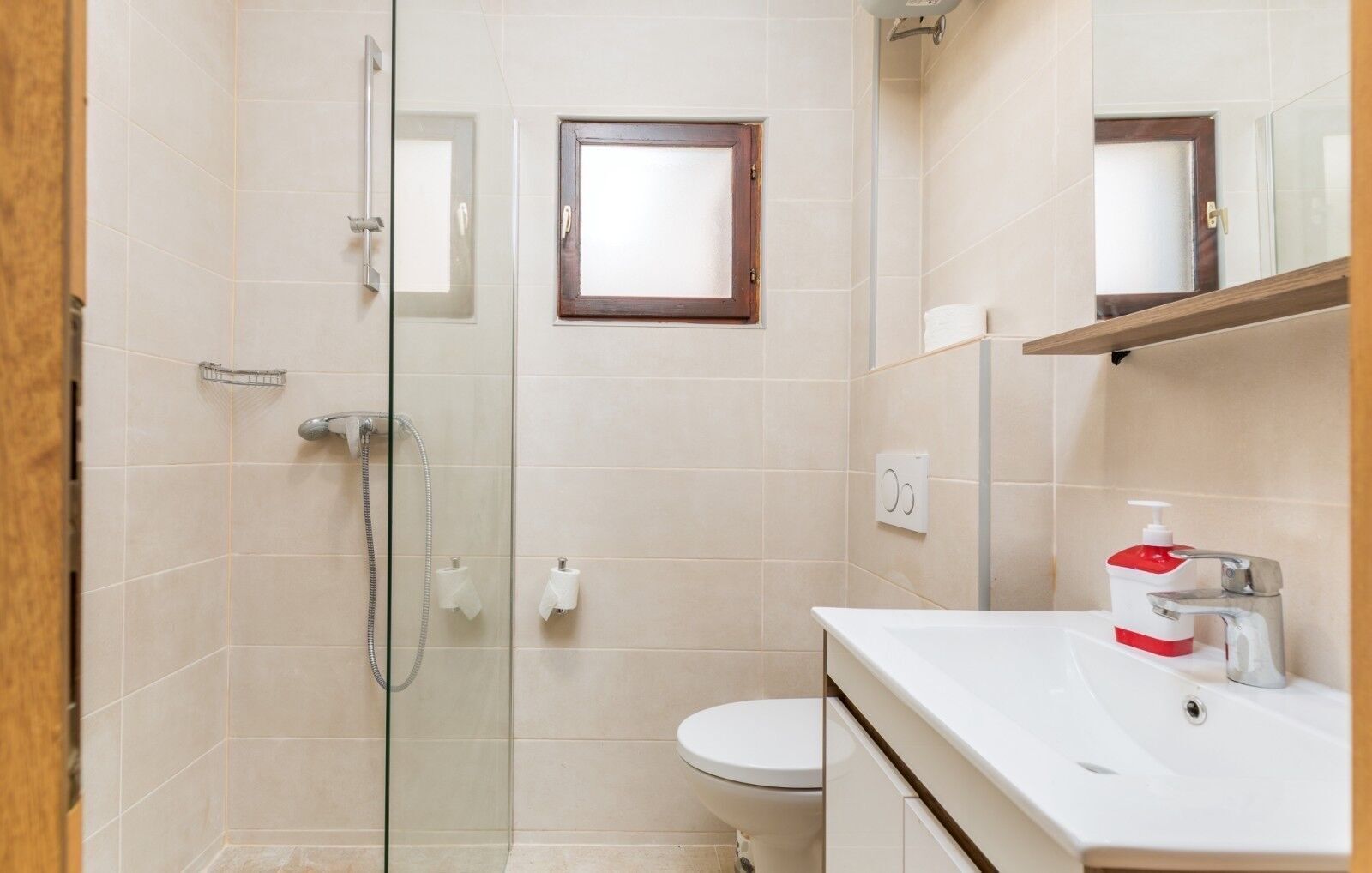 A modern bathroom with a glass shower enclosure, a toilet, and a white sink with a red soap dispenser.