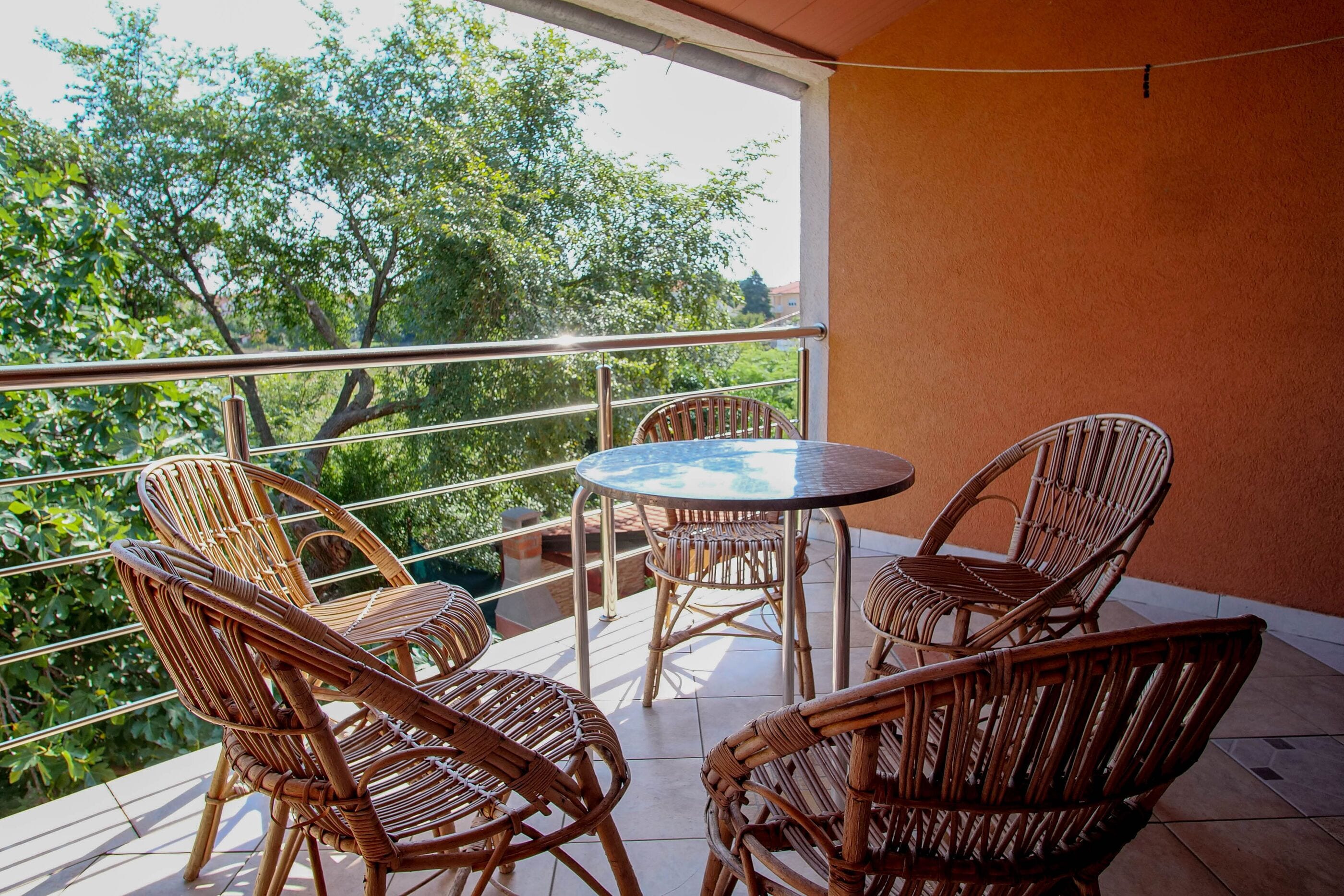 Un balcon avec des fauteuils en osier et une table ronde, donnant sur un jardin arboré.