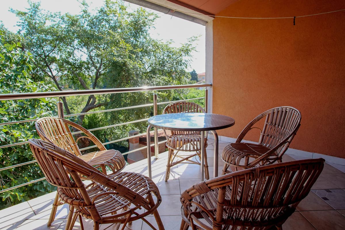 Un balcon avec des fauteuils en osier et une table ronde, donnant sur un jardin arboré.