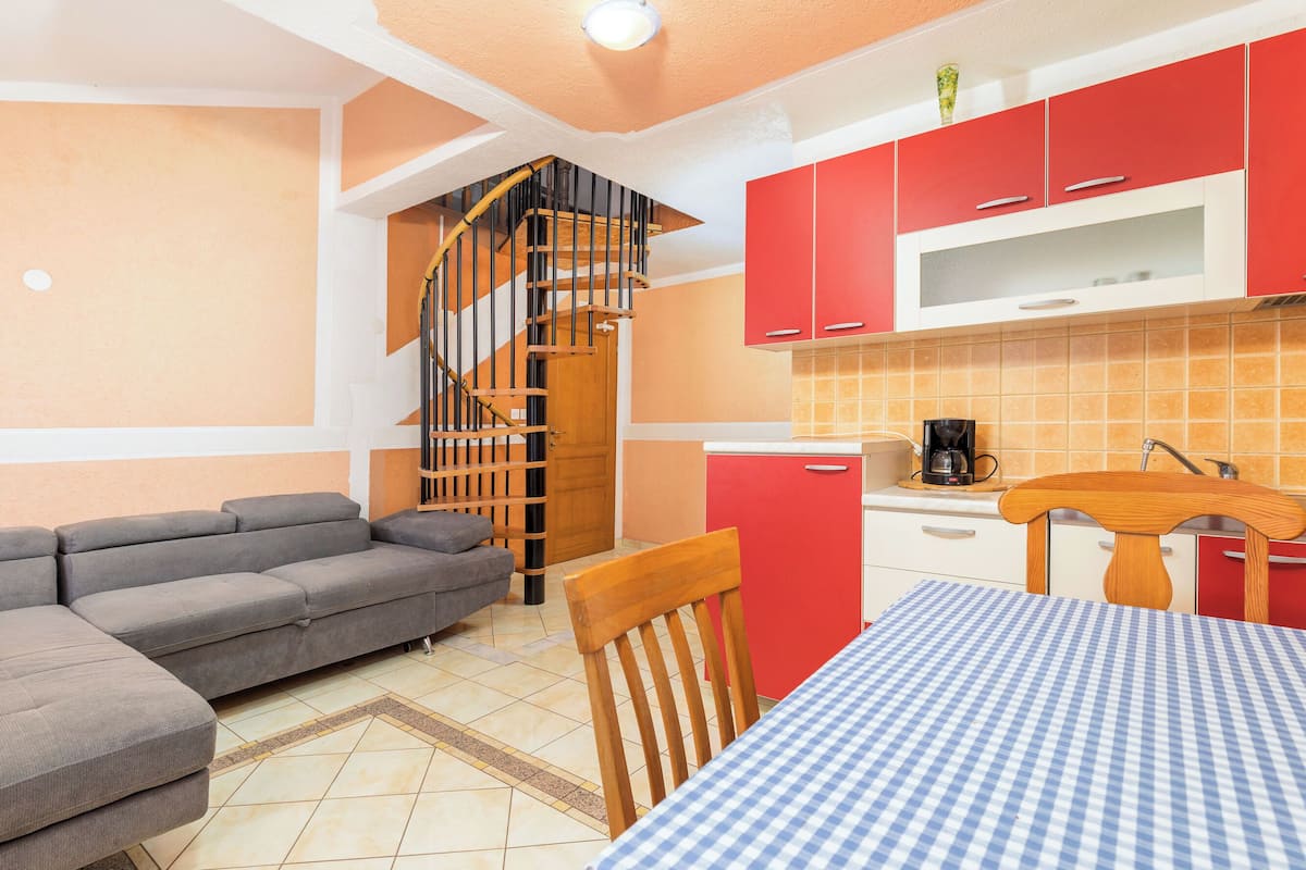 Un espace de vie compact comprenant un canapé d’angle gris, une table de salle à manger en bois recouverte d’une nappe à carreaux, une cuisine rouge dotée d’un four à micro-ondes et d’une machine à café, ainsi qu’un escalier en colimaçon.