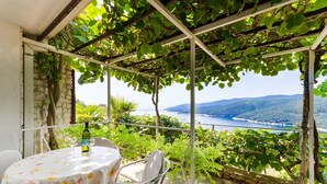 A covered outdoor seating area with a table and chairs, overlooking a scenic view of a body of water and hills. - Apartments Mandica (Rabac)