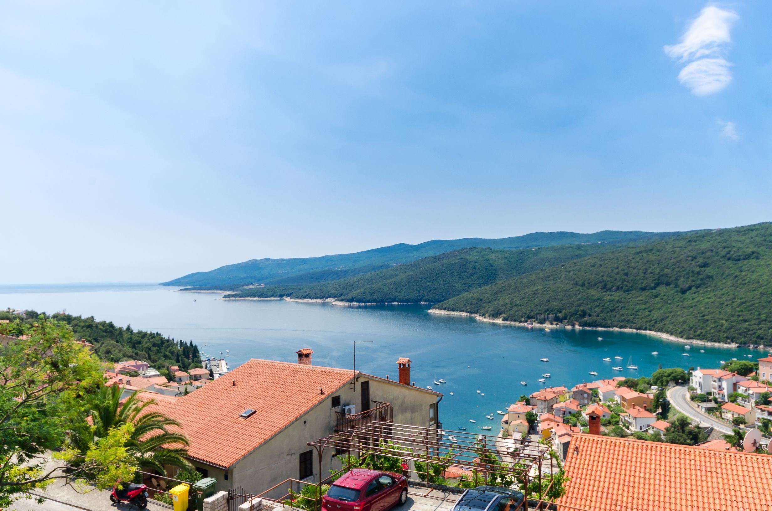 A coastal town with terracotta-roofed buildings, a marina with boats, and a clear view of the sea and distant mountains.