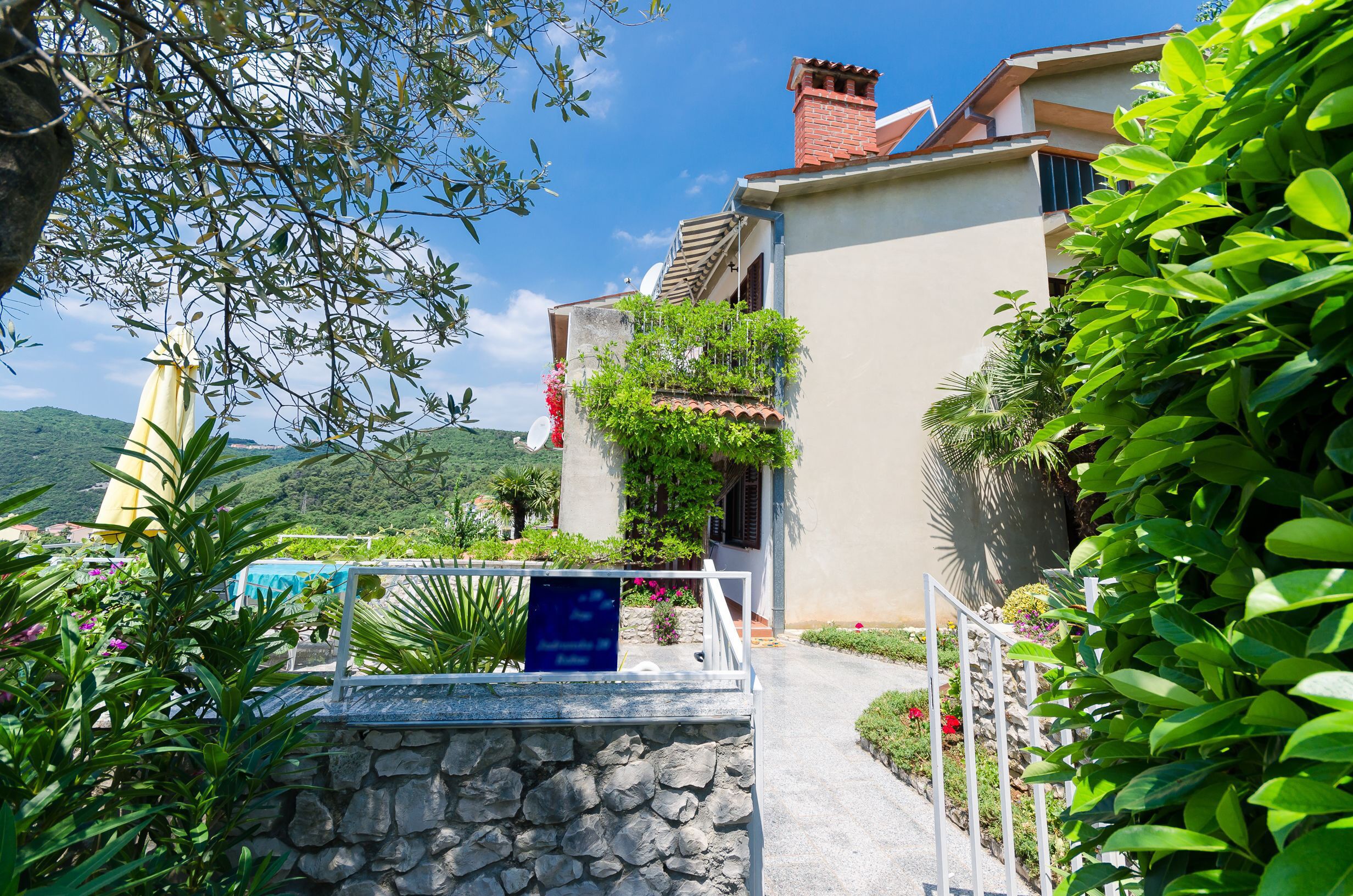 Une villa avec un chemin de pierre menant à un portail blanc, entourée d’une végétation luxuriante et sous un ciel d’un bleu limpide.