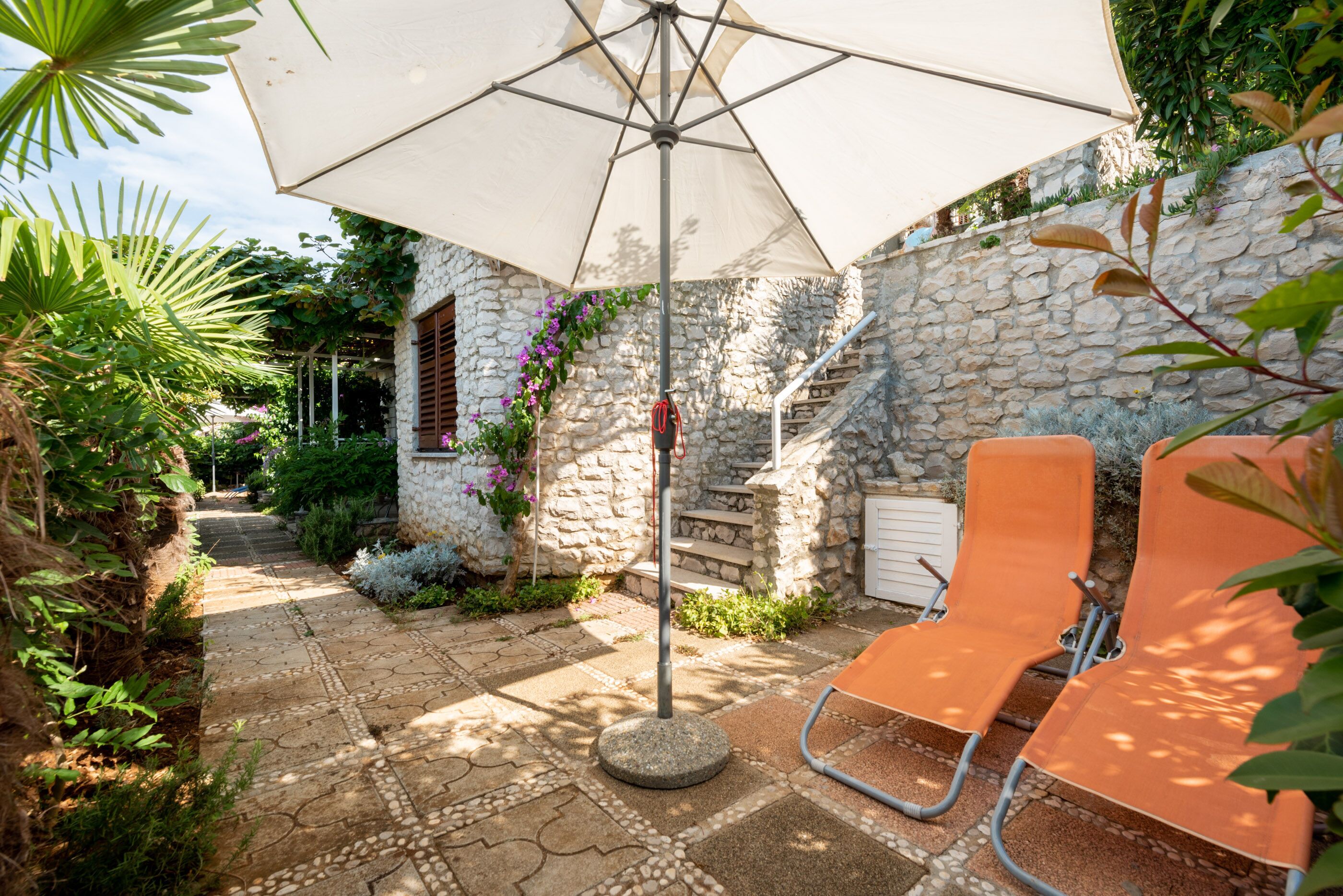 A stone-walled patio with a tiled pathway, outdoor seating, and a large umbrella.
