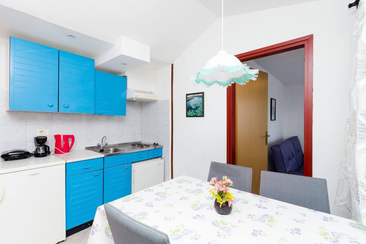 A compact kitchen with blue cabinets, a white countertop, and a dining area with a table set for two.