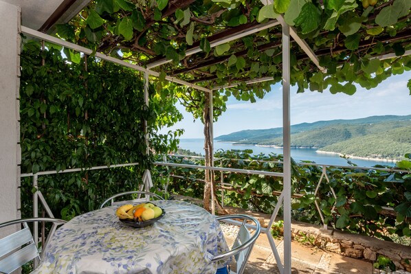 A covered outdoor seating area with a table set for two, surrounded by lush greenery and a scenic view of a coastal landscape. - Apartments Mandica (Rabac)