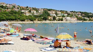 A beach with people sunbathing and swimming, surrounded by hillside buildings. - Apartments Mandica (Rabac)