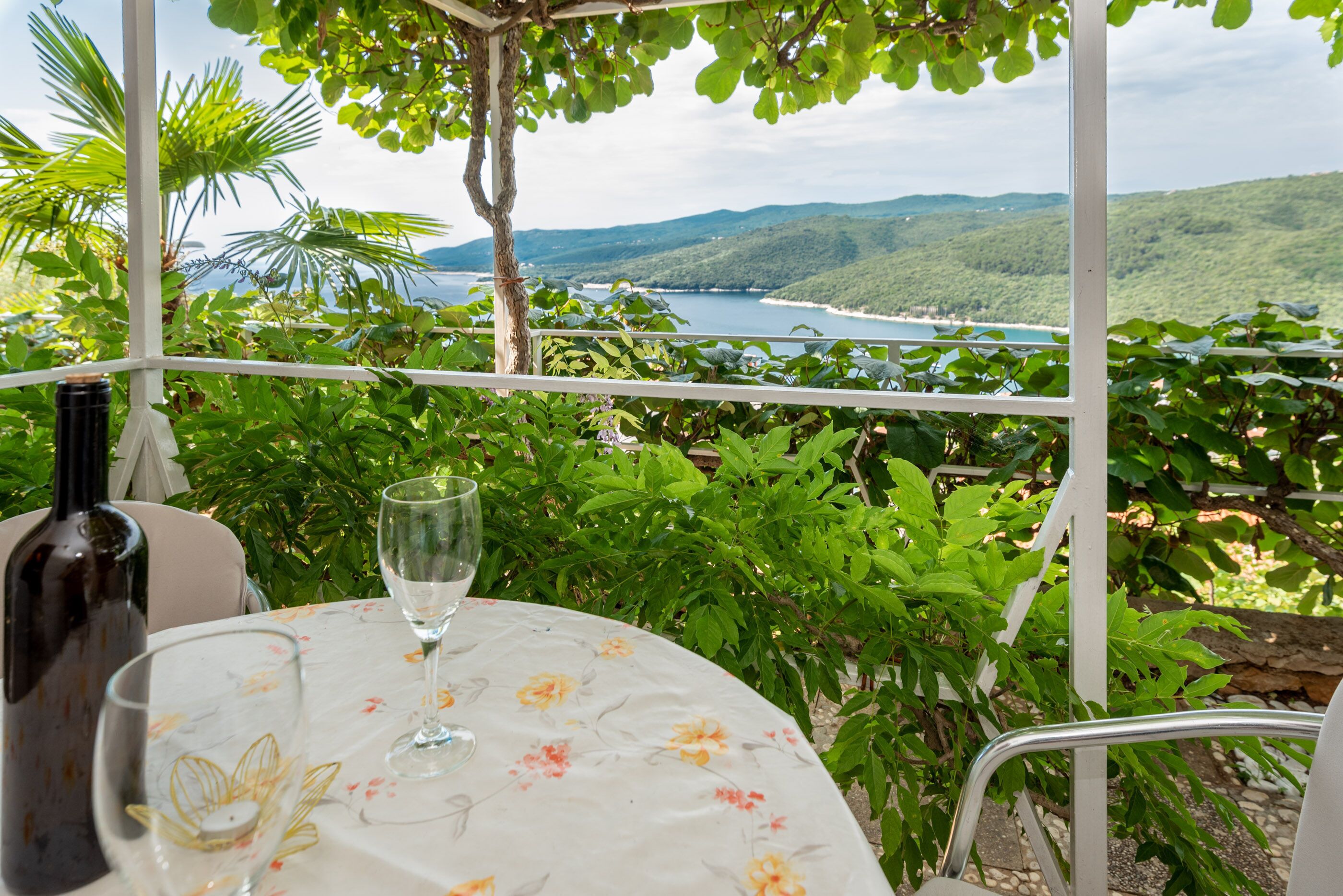 A table with a bottle and a glass, set against a backdrop of lush greenery and a coastal view.