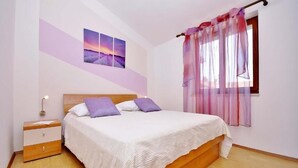 A bedroom with a wooden bed, a nightstand, a wall-mounted lamp, and a window with sheer curtains. - House Bruno (Pula)