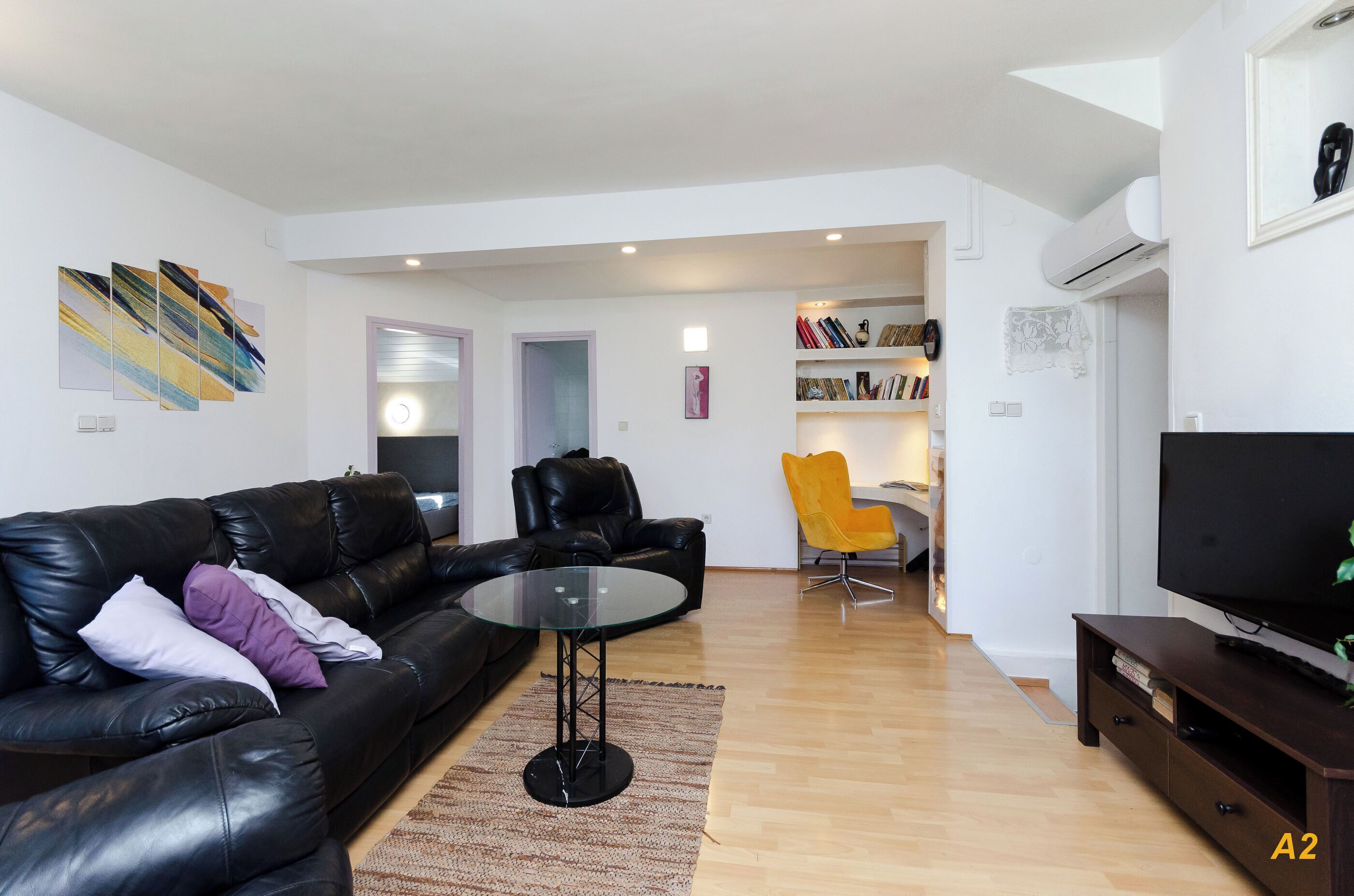 A modern living room with a black leather sofa, a glass coffee table, a yellow chair, a flat-screen TV, and a bookshelf.
