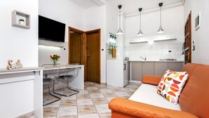 Een moderne keuken met witte kasten, een eethoek met een witte tafel en stoelen, en een oranje bank met een kleurrijk kussen.