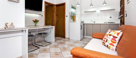 A modern kitchen with white cabinets, a dining area with a white table and chairs, and an orange sofa with a colorful pillow.