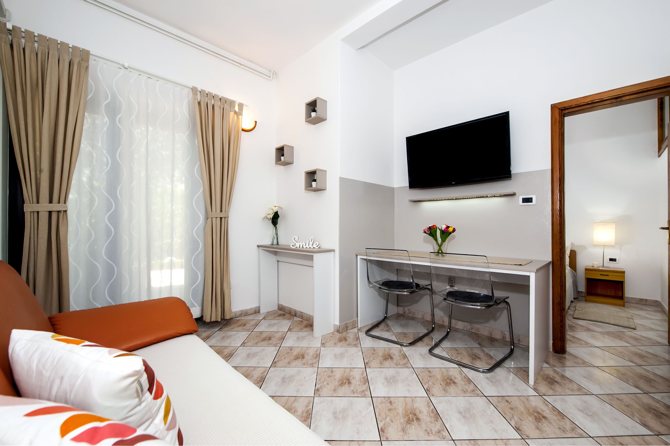 A modern hotel room with a sofa, a desk with a chair, a TV mounted on the wall, and a small bedside table with a lamp.