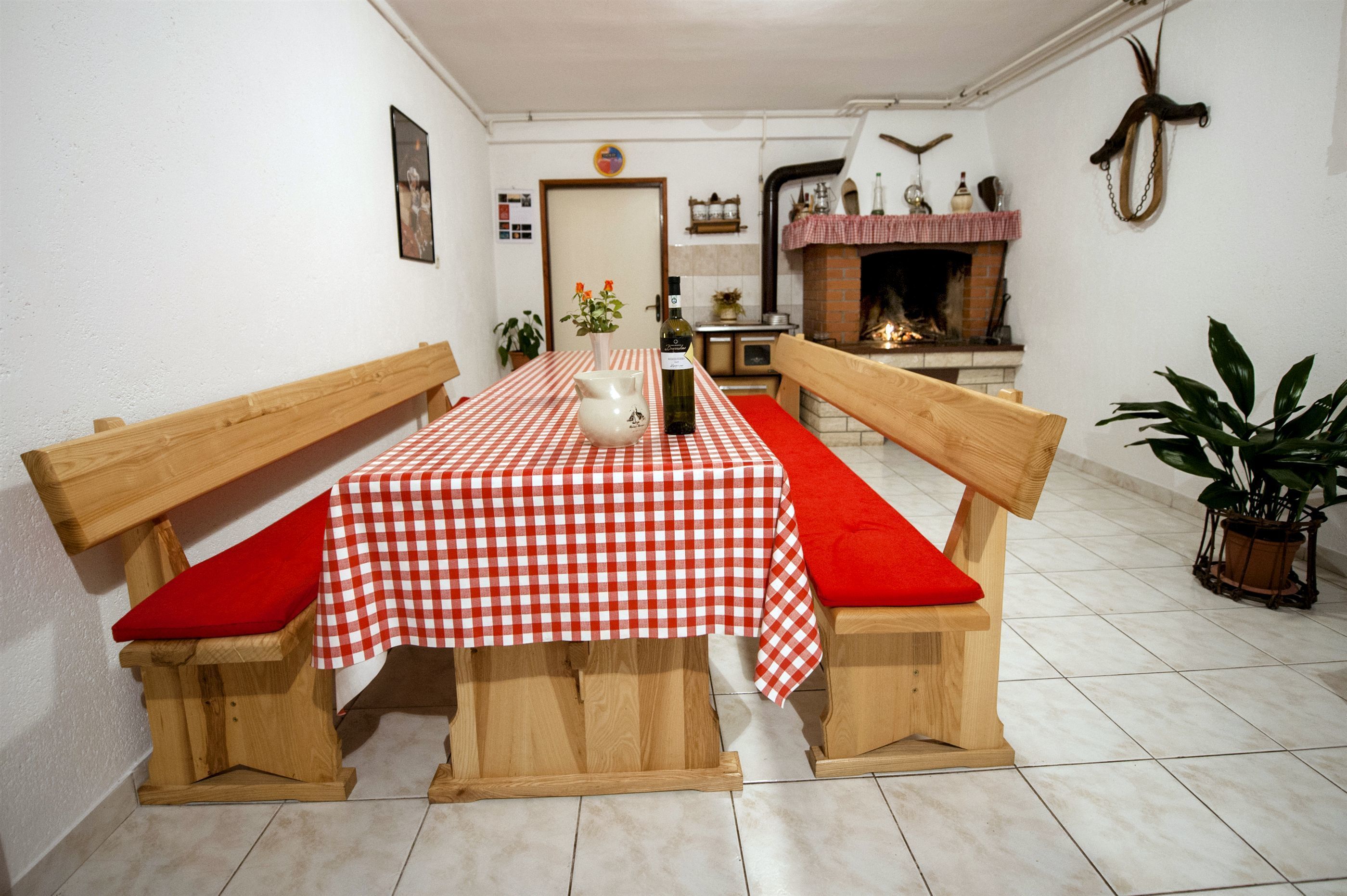 A dining area with a checkered tablecloth, wooden benches with red cushions, a vase, and a bottle of wine.