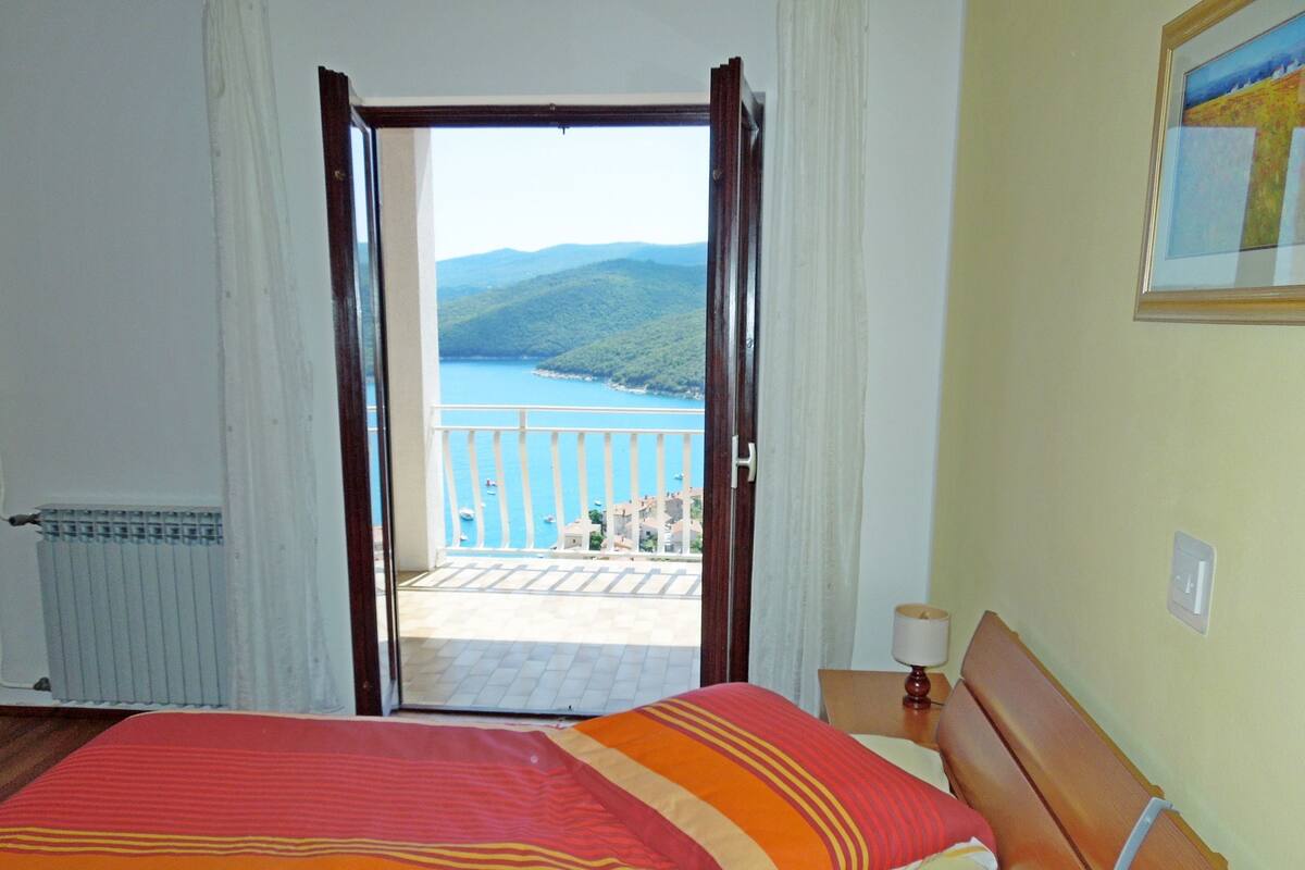 A bedroom with a bed, a nightstand, a lamp, a window with a view of a coastal landscape, and a painting on the wall.