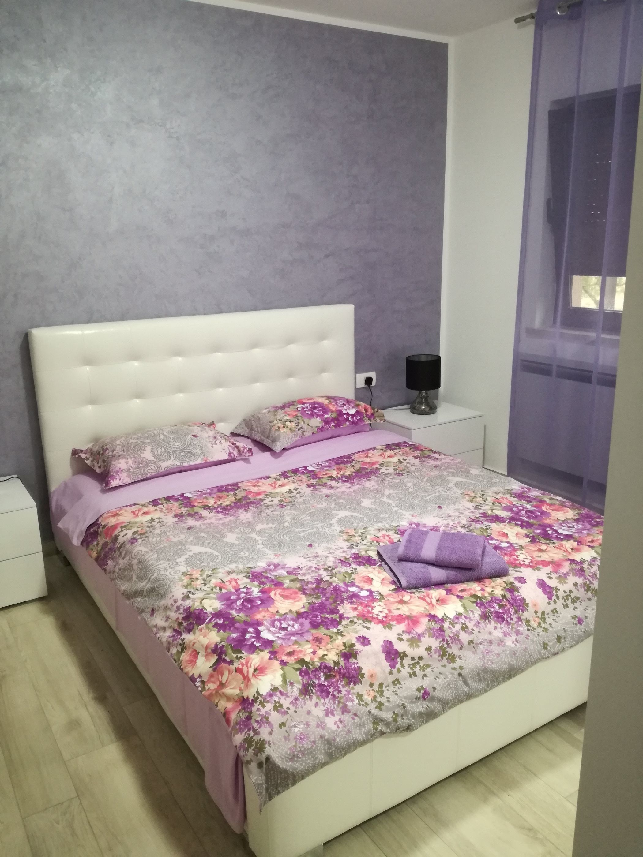 A bedroom with a white bed, floral bedding, a nightstand with a lamp, and a window with purple curtains.