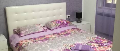 A bedroom with a white bed, floral bedding, a nightstand with a lamp, and a window with purple curtains.