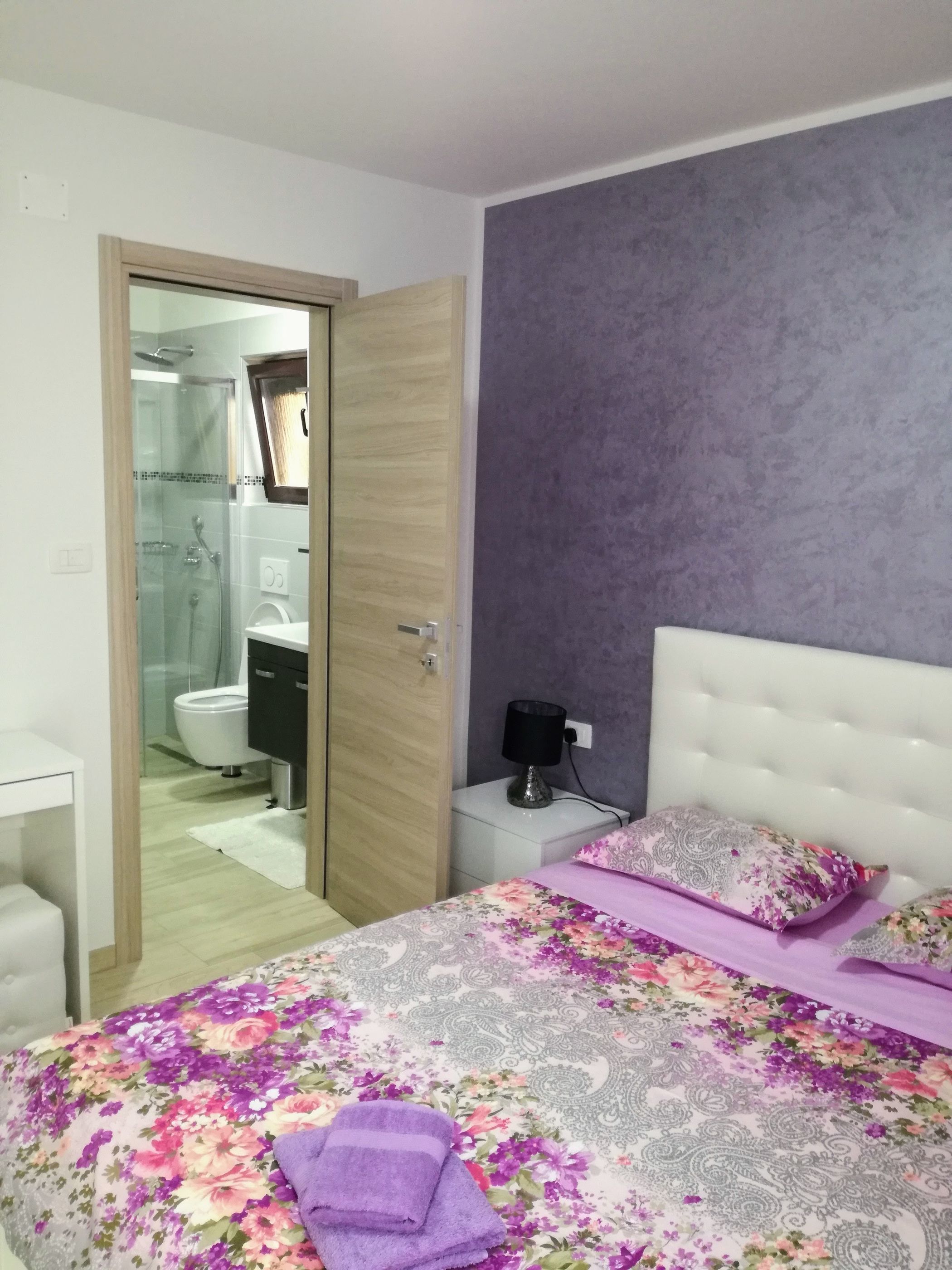 A bedroom with a bed, a nightstand, a bedside table, a lamp, and a bathroom visible through an open door.