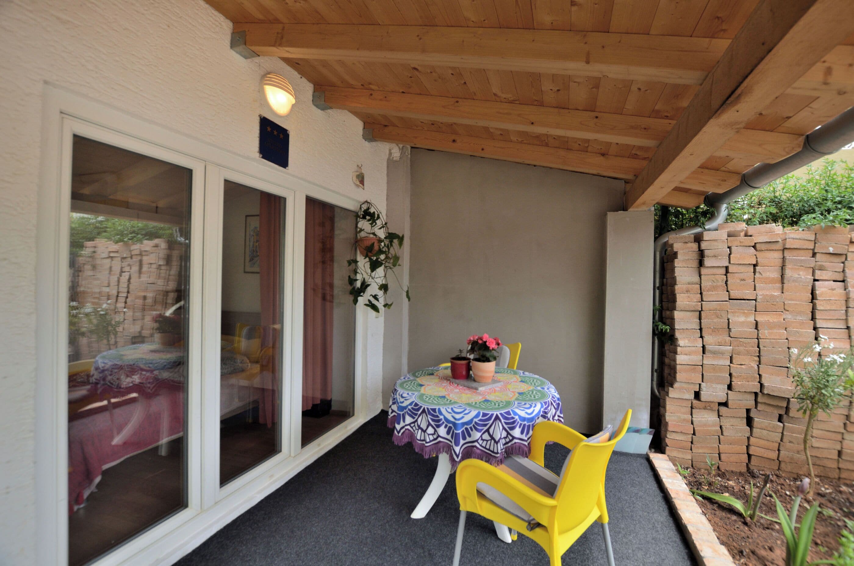 A covered patio with a round table, yellow chairs, and a brick wall.