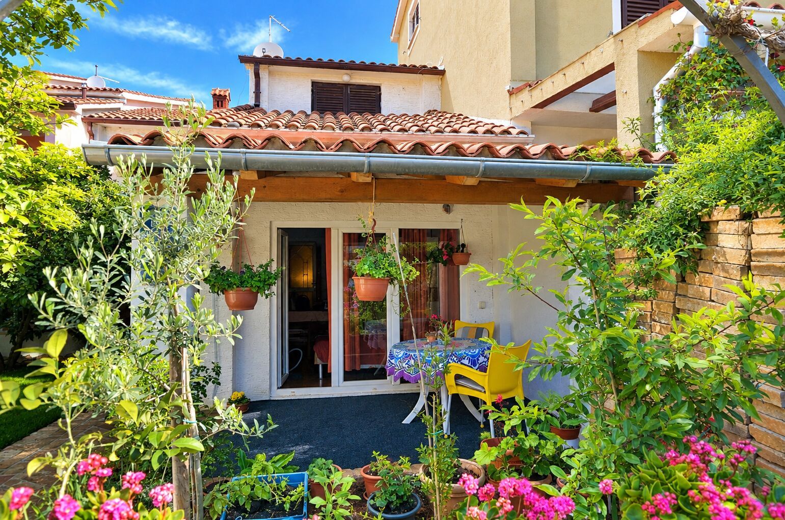 A house with a tiled roof, a covered patio with a table and chairs, and various potted plants.