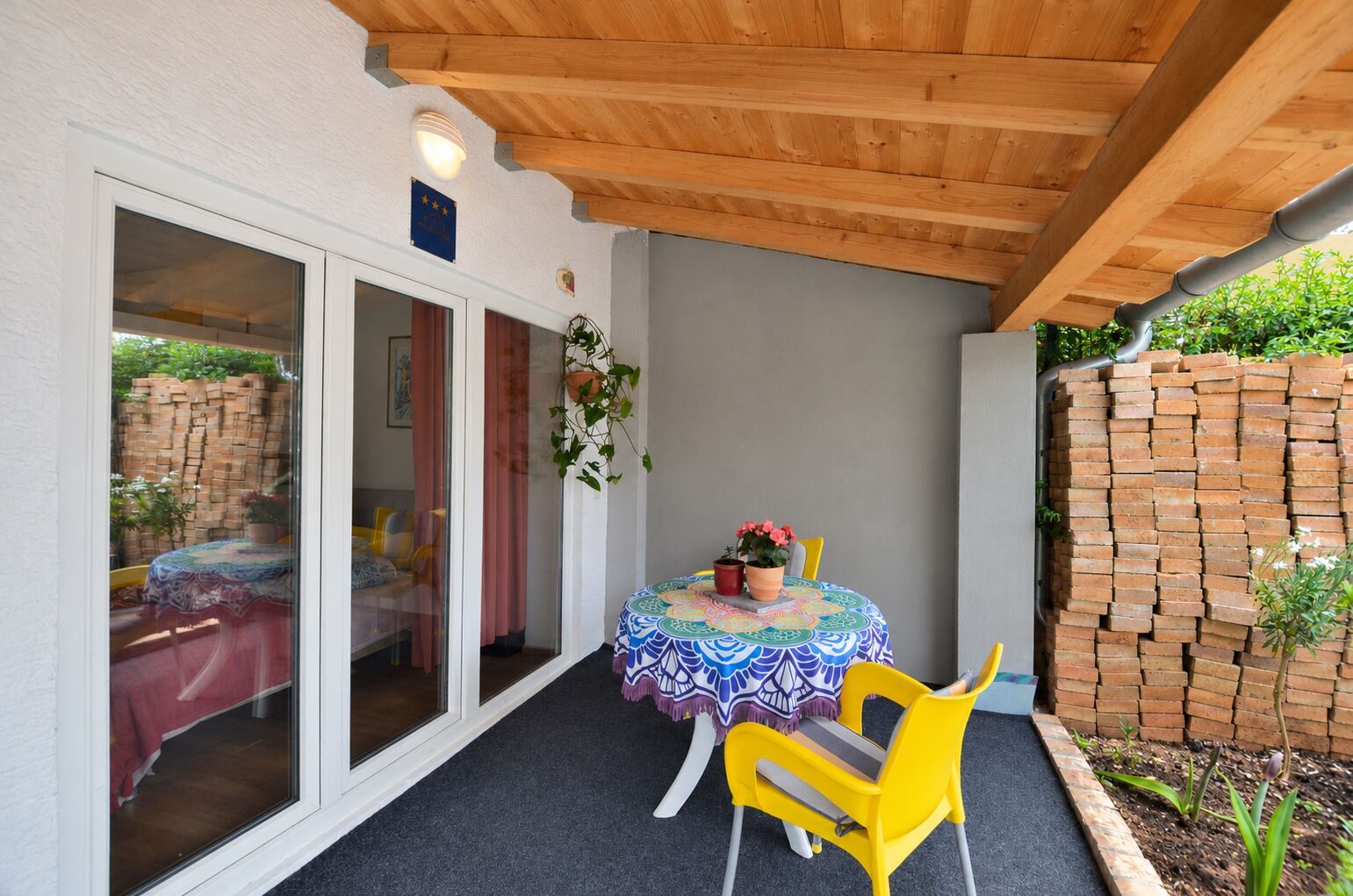 Un patio techado con una mesa redonda, sillas amarillas y una pared de ladrillo.