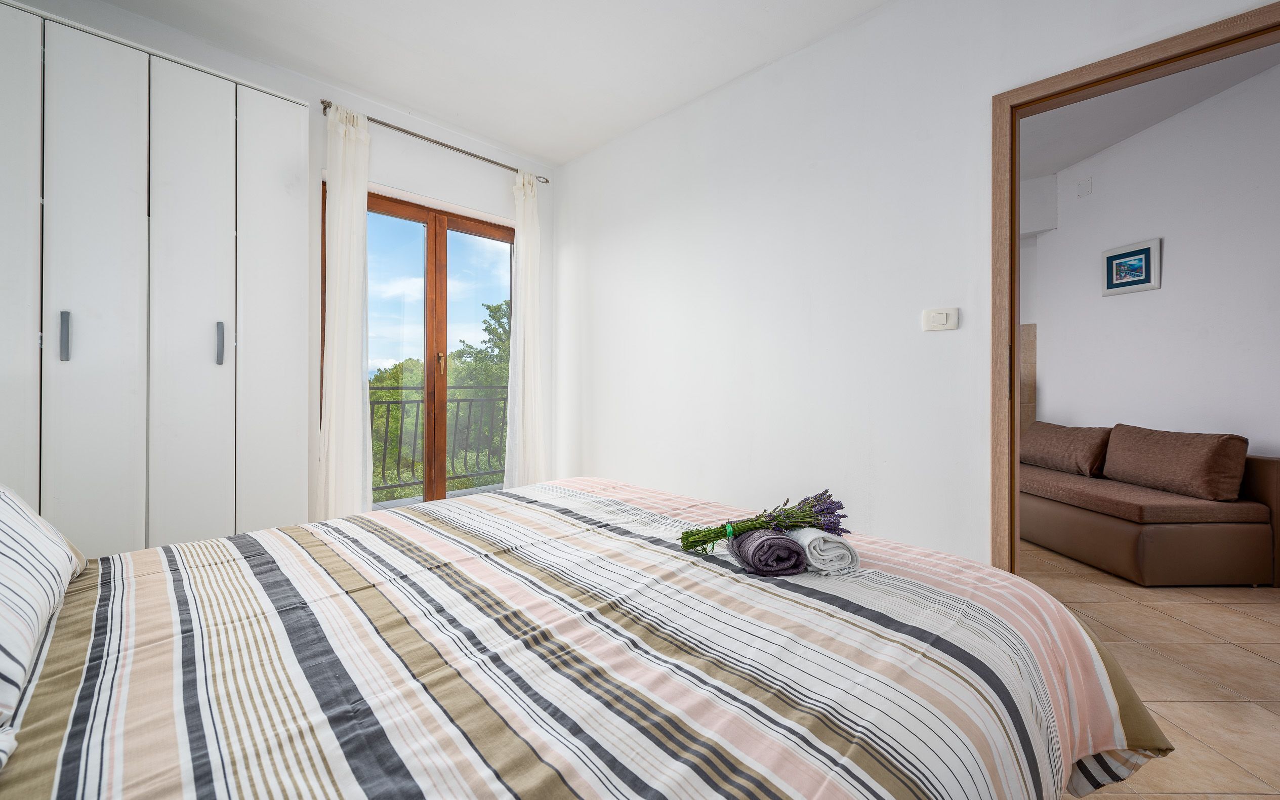 A bedroom with a bed, a wardrobe, a balcony with a view, and a brown sofa.