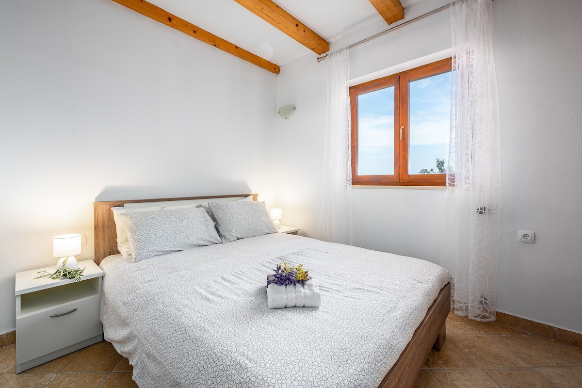 A bedroom with a bed, nightstands, a window with curtains, and a wooden ceiling beam.