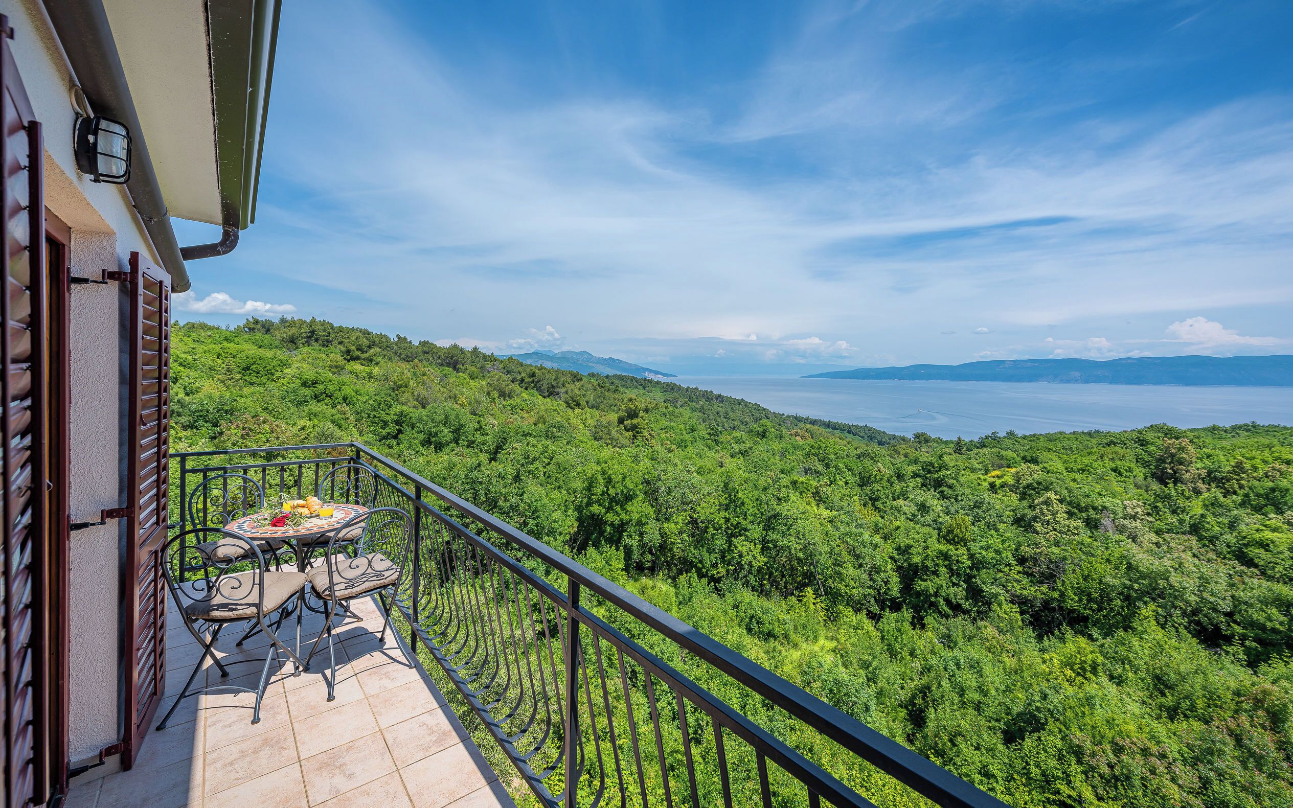 A balcony with a table and chairs, overlooking a lush green landscape and a body of water.