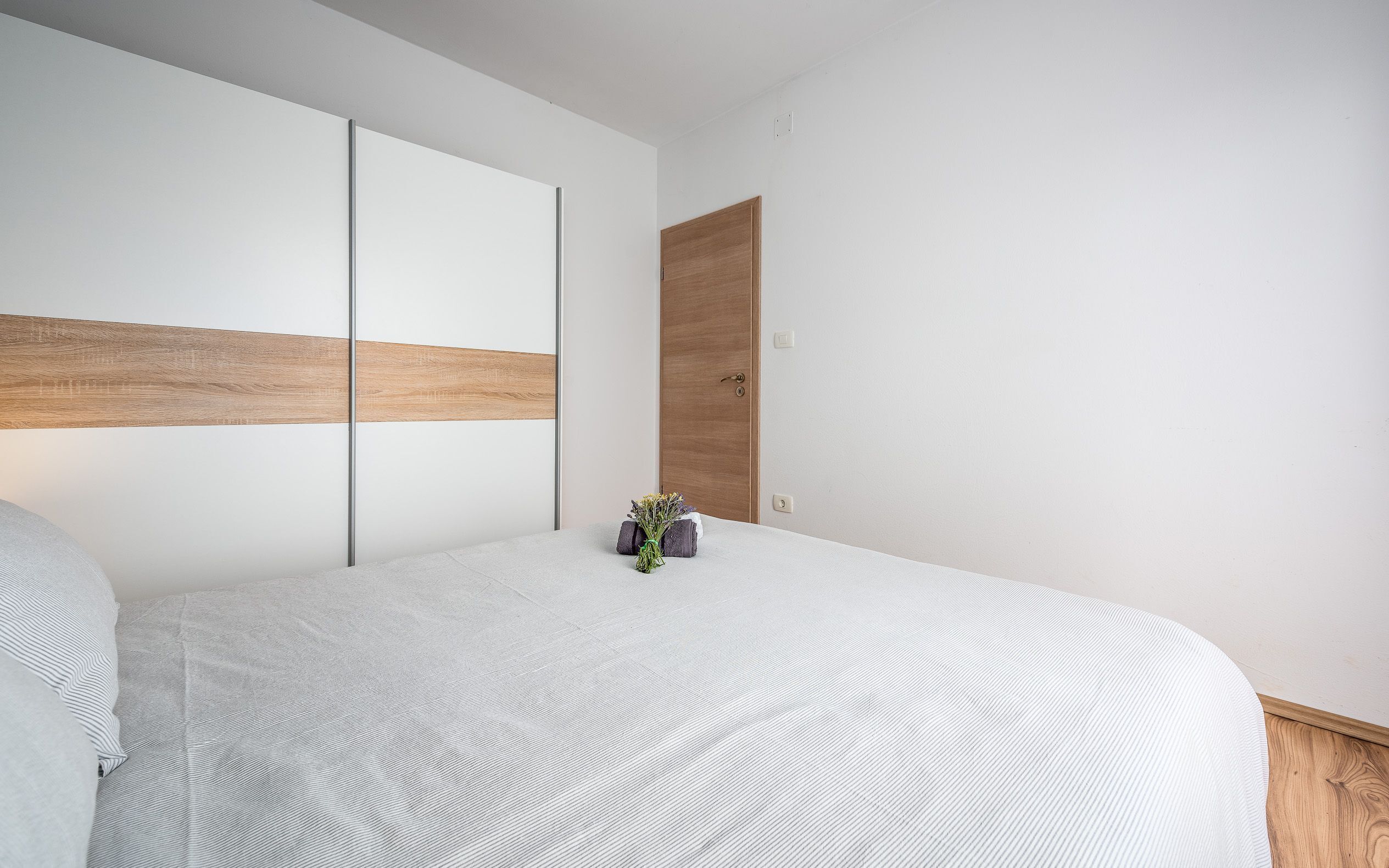 A minimalist bedroom with a large bed, a wooden door, and a small vase of flowers on the nightstand.