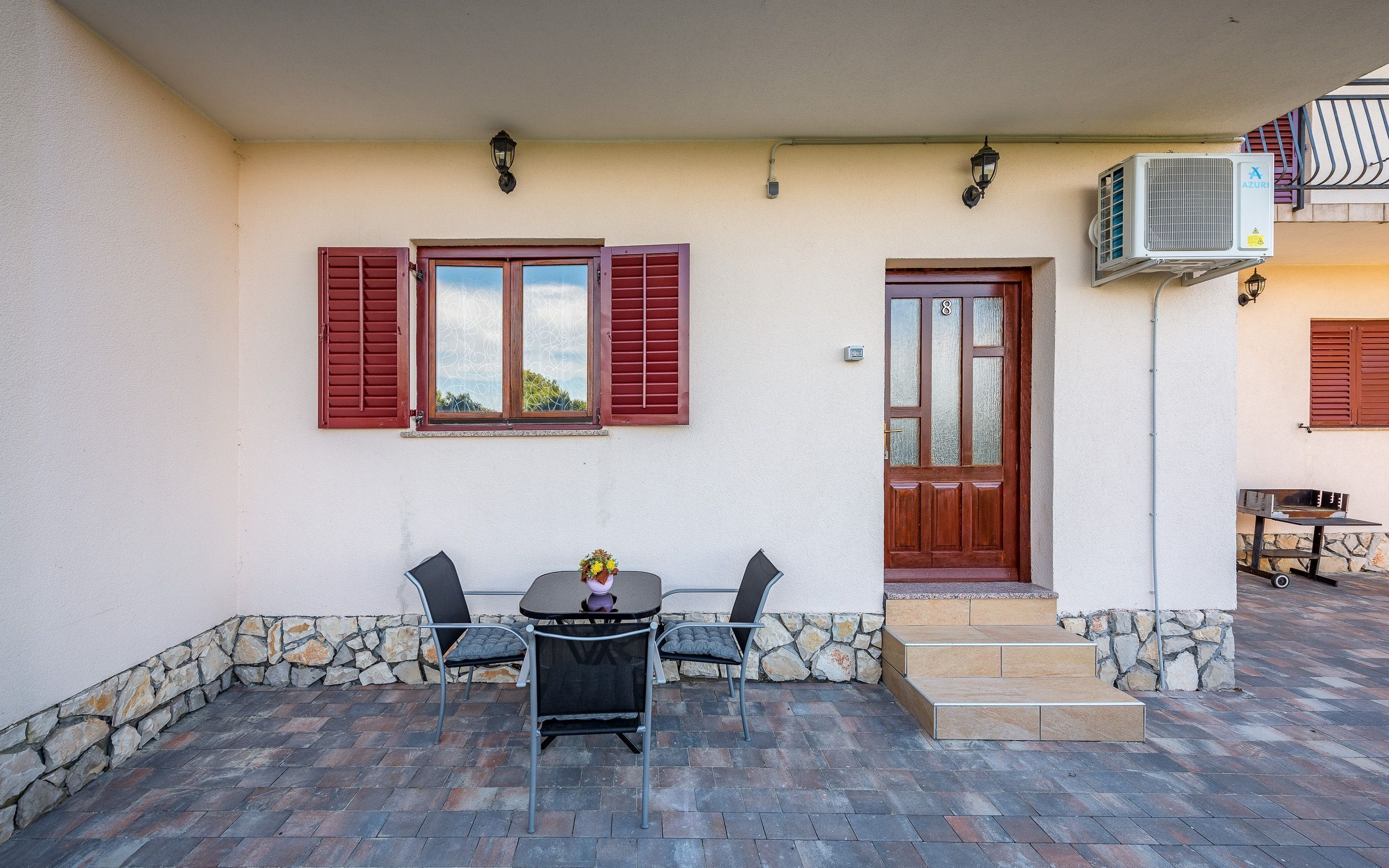 A building with a stone wall foundation, a wooden door, and a window with shutters. There is a table and two chairs outside.
