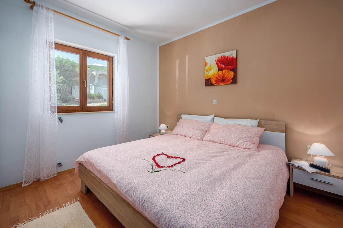 A bedroom with a bed, a window with curtains, a nightstand, a lamp, and a painting on the wall.